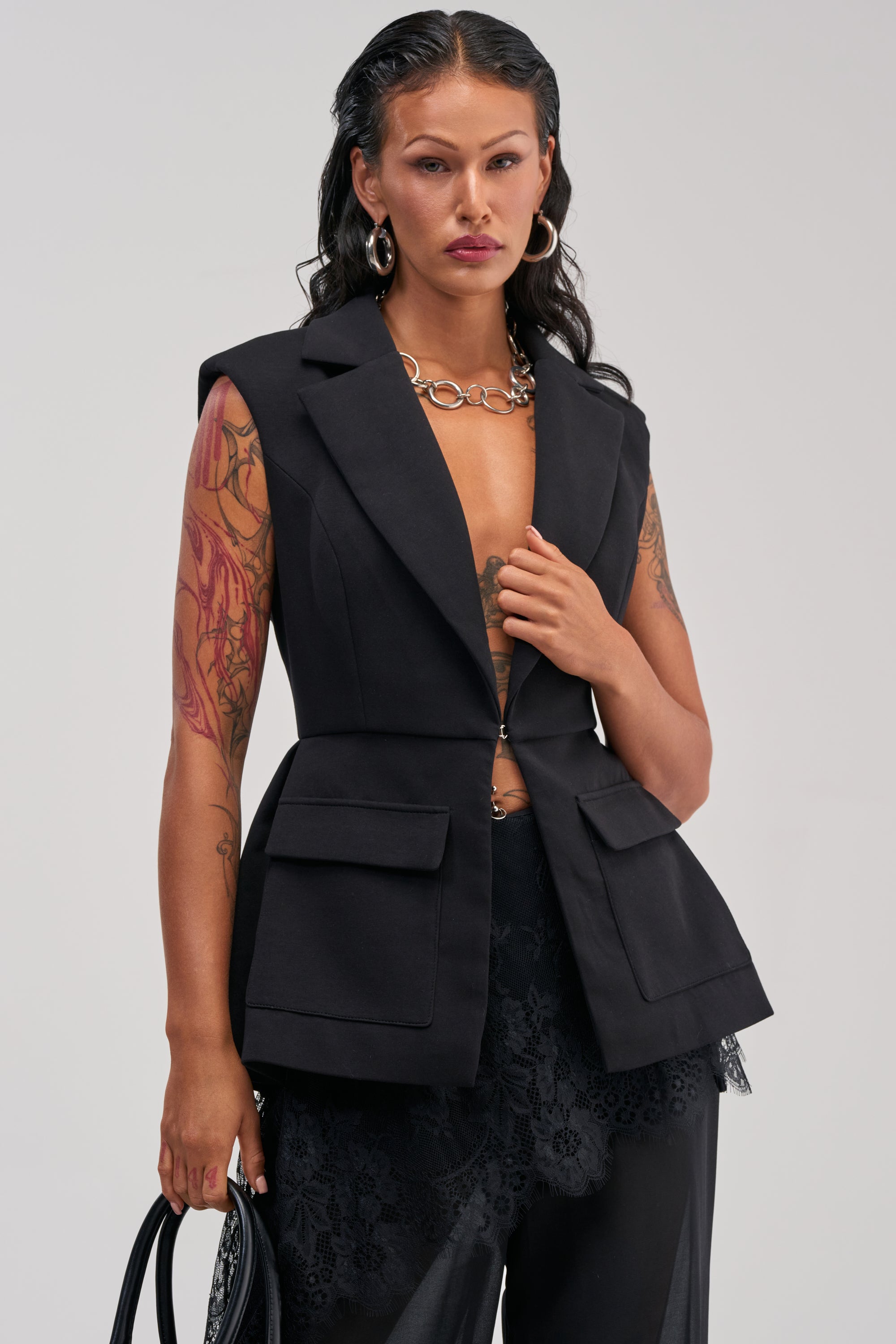 A person with long dark hair, tattoos, and hoop earrings wears the NAOMI BLAZER VEST IN BLACK and lace pants, holding a black bag, standing against a plain light background.