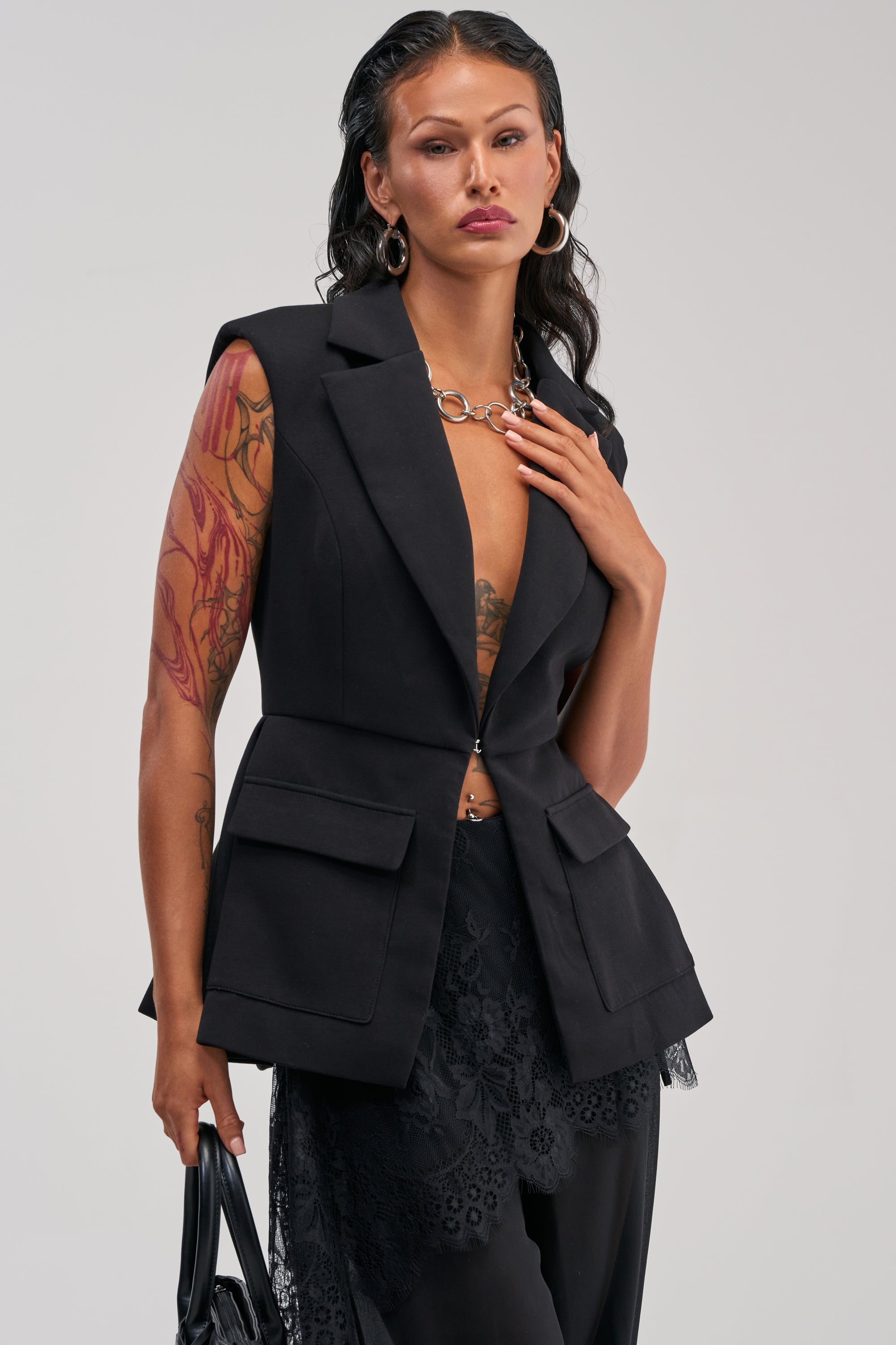 A woman with long dark hair wears the NAOMI BLAZER VEST IN BLACK, styled with black lace pants that reveal her arm tattoos. She accessorizes with a black handbag and a silver chain necklace, posing confidently against a plain background for an elevated twist on tailored office attire.