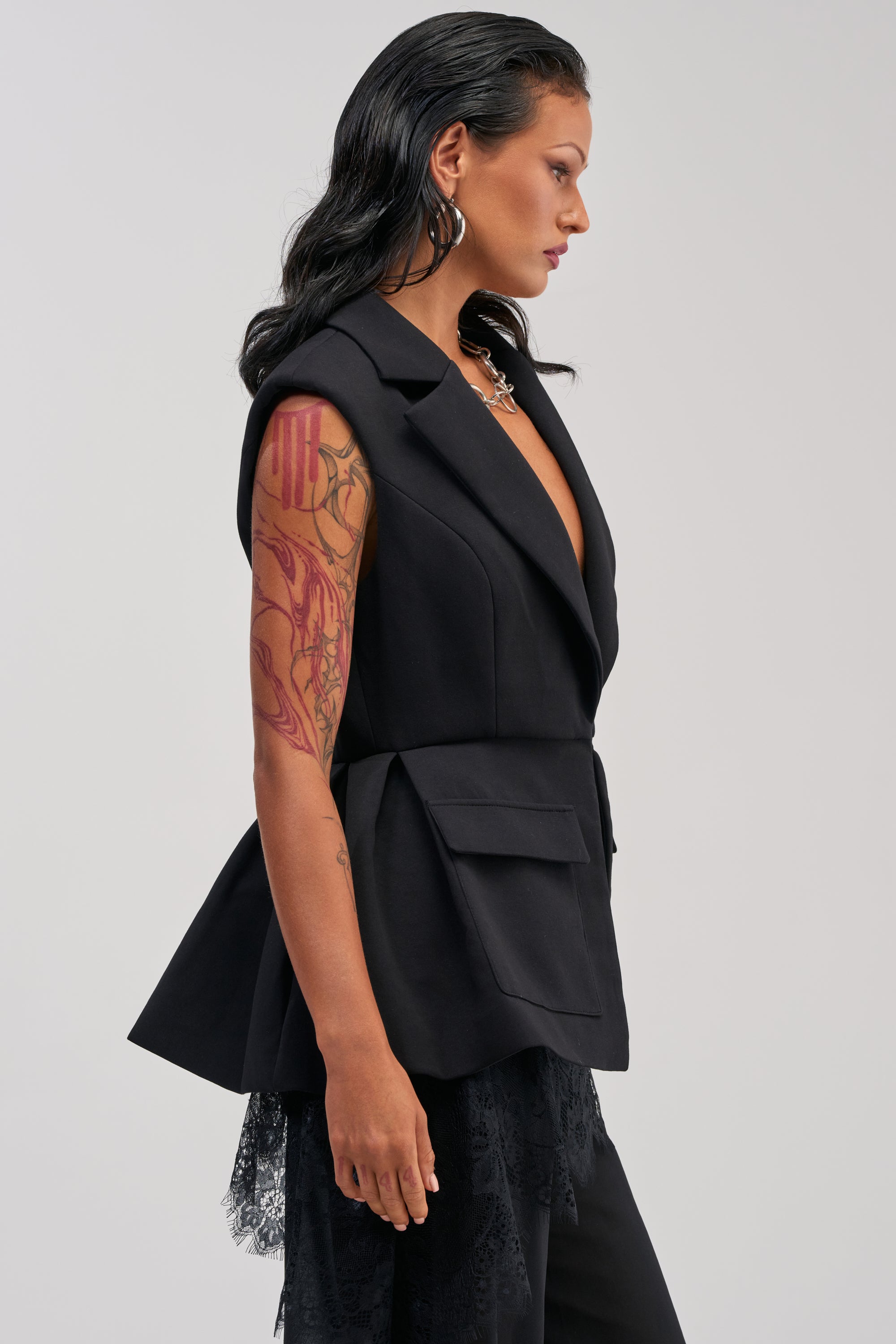Wearing the NAOMI BLAZER VEST IN BLACK, a woman with long dark hair and arm tattoos stands in profile against a plain background. The modern, sleeveless blazer vest features large pockets and lace detailing—a fresh take on tailored office wear.
