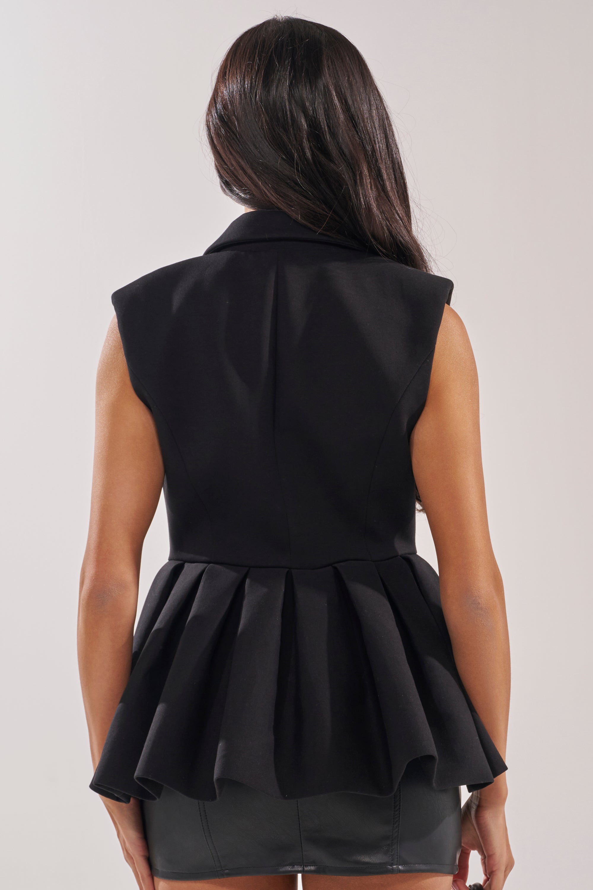 A woman is seen from the back wearing the NAOMI BLAZER VEST IN BLACK, a sleeveless, tailored piece with a peplum hem, paired with a black skirt. Her long dark hair flows down, creating an elegant office look against a light background. 3