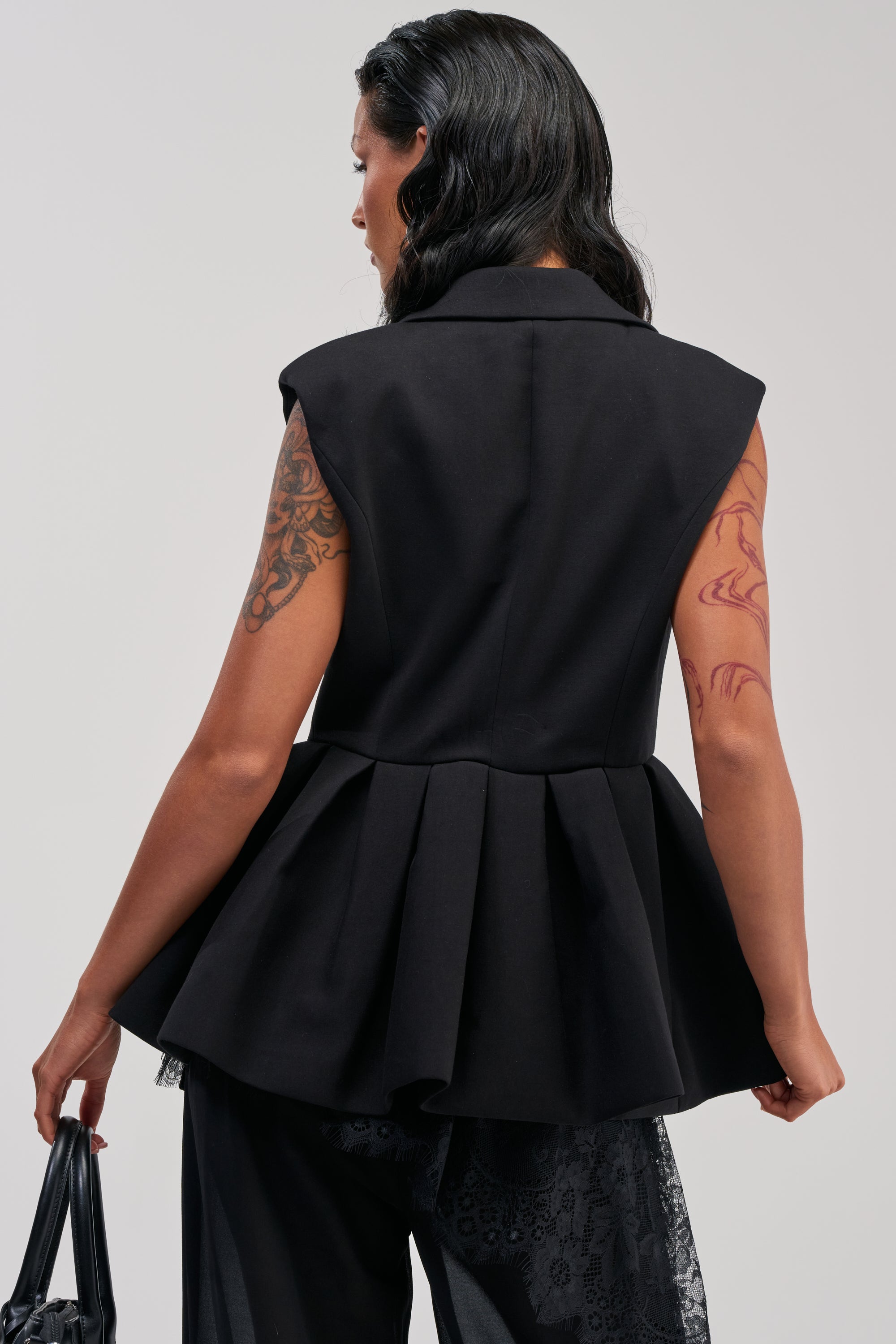 A woman with long dark hair and visible tattoos stands facing away, holding a black bag against a light gray background, modeling the NAOMI BLAZER VEST IN BLACK—a modern sleeveless blazer style perfect for tailored office wear.