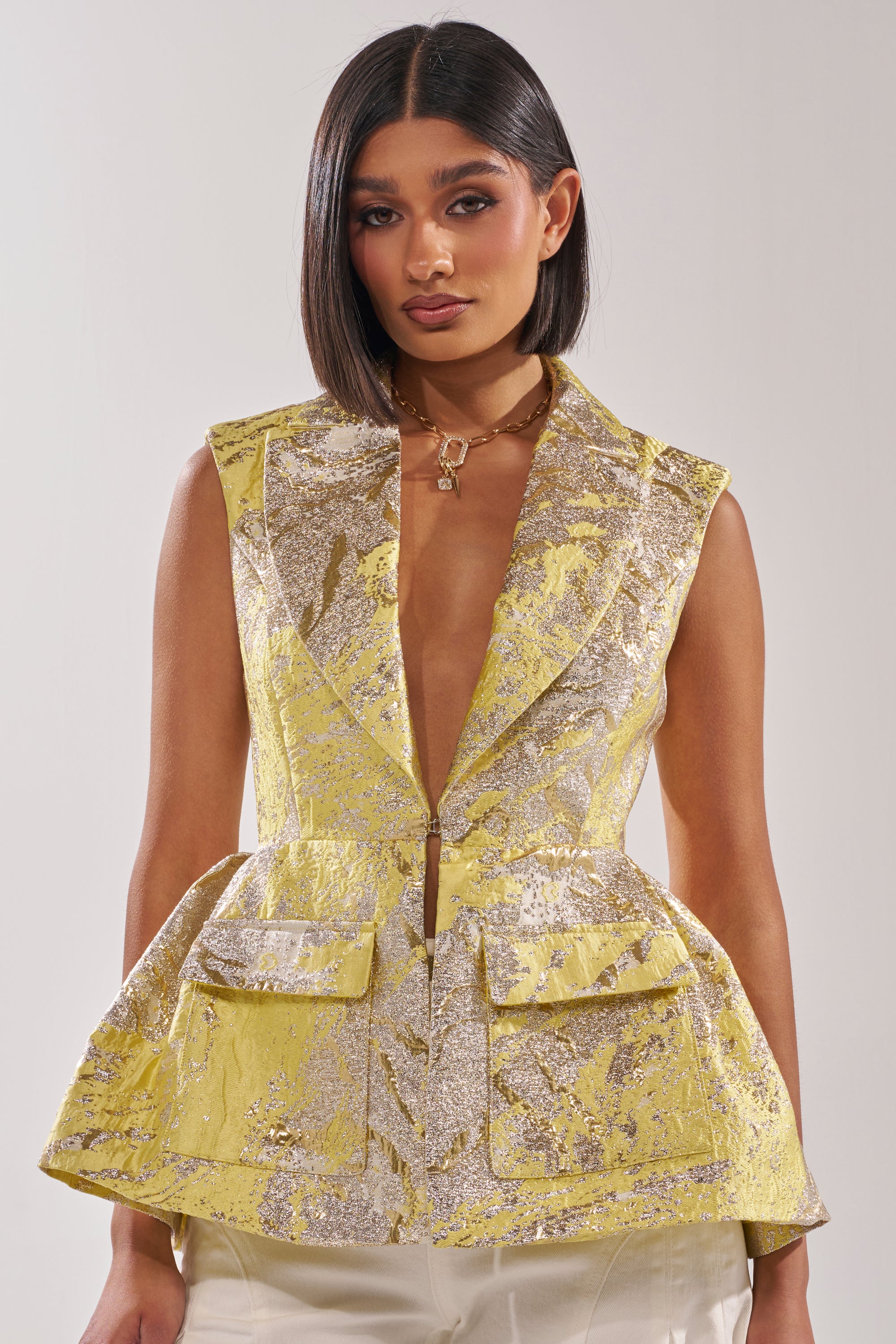 A woman with straight shoulder-length hair models the NAOMI BLAZER VEST IN BROCADE, a yellow metallic sleeveless vest with a deep V neckline, peplum hem, and front pockets, paired with cream pants against a plain background. 0