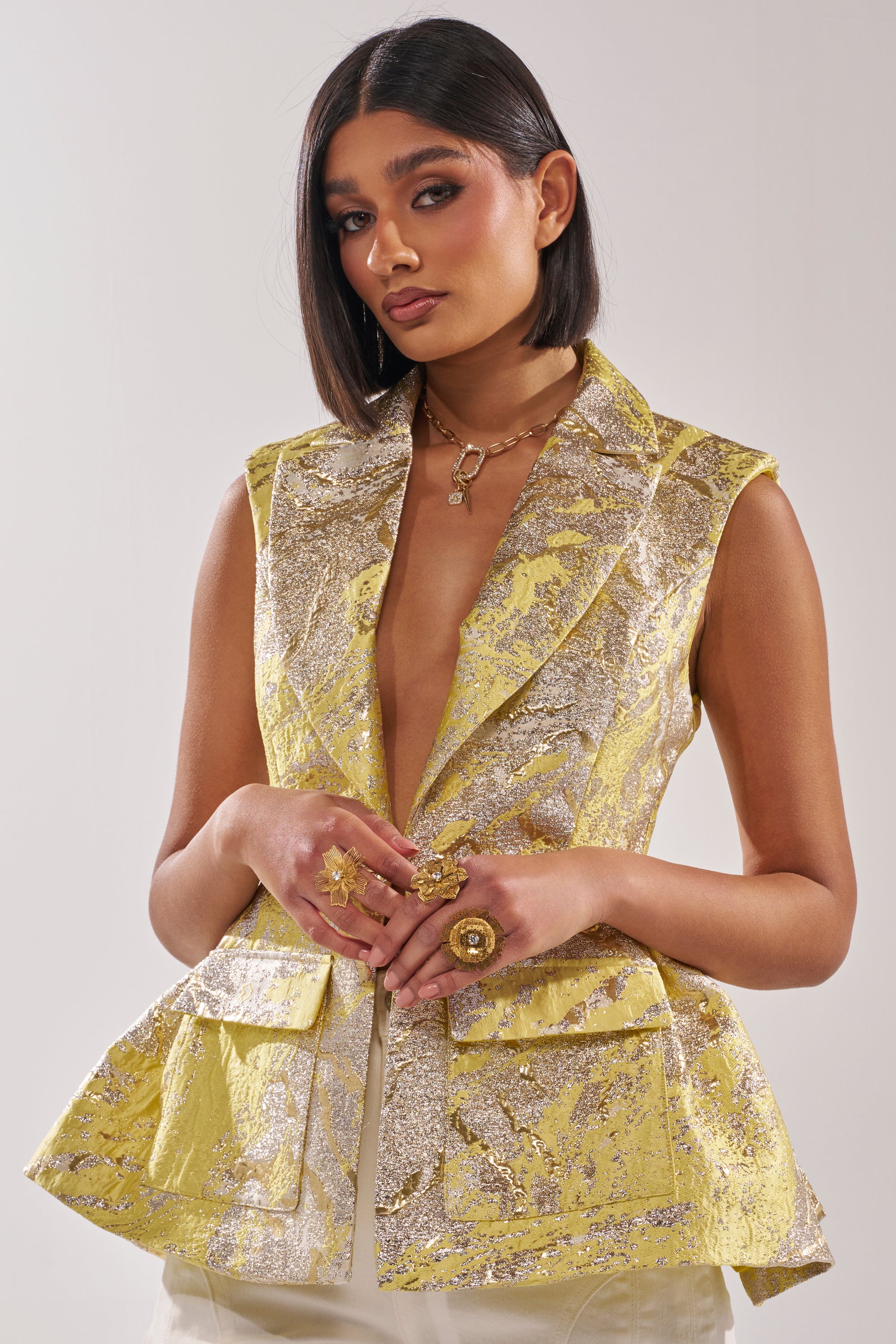 A woman with straight, shoulder-length dark hair models the NAOMI BLAZER VEST IN BROCADE, paired with chunky gold rings and a gold necklace, against a plain light background. 1
