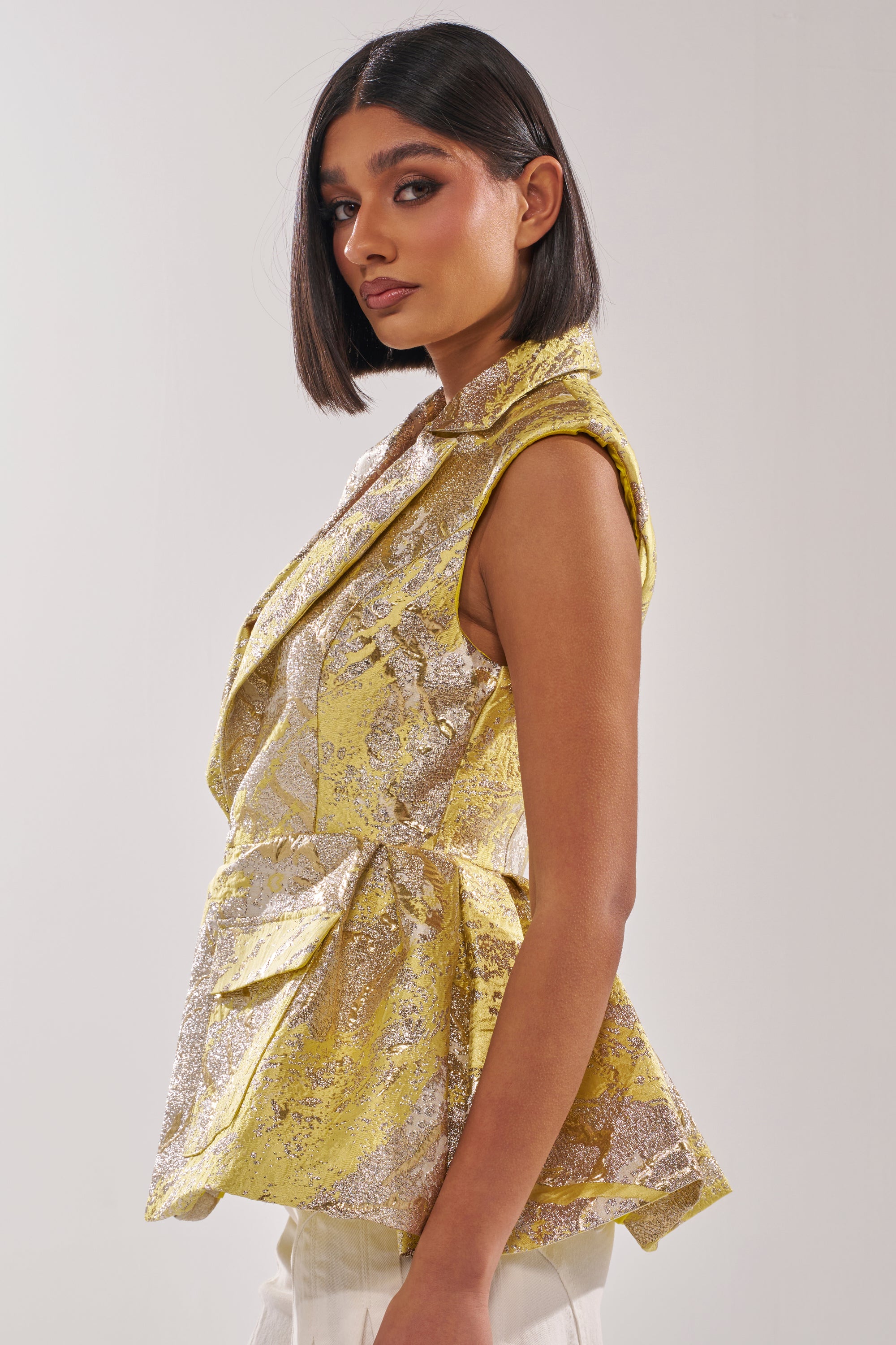 A woman with straight, shoulder-length dark hair wears the NAOMI BLAZER VEST IN BROCADE, styled sleeveless in gold and silver, paired with white pants, shown in profile against a plain light background. 2