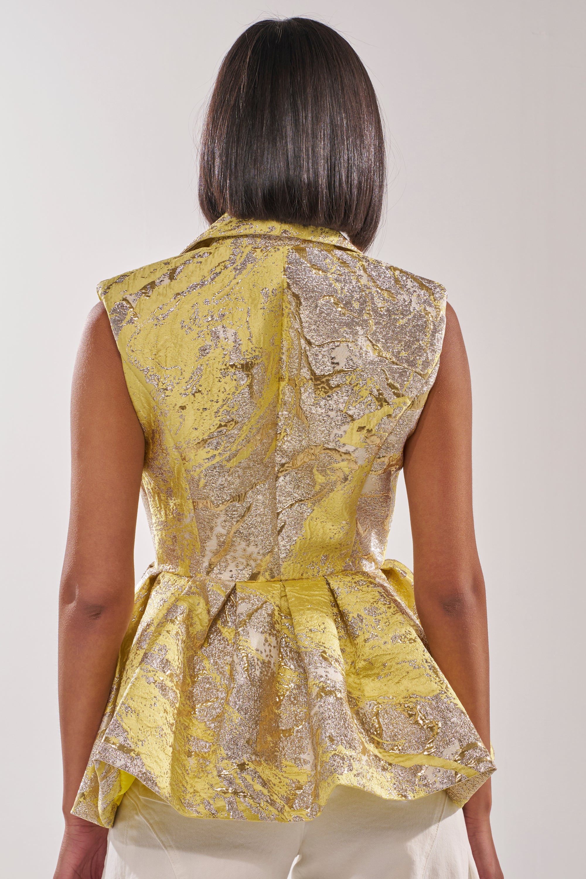A woman with straight, dark hair is seen from the back wearing the NAOMI BLAZER VEST IN BROCADE, a sleeveless yellow metallic peplum vest with a flared waist, paired with cream pants against a light background. 3