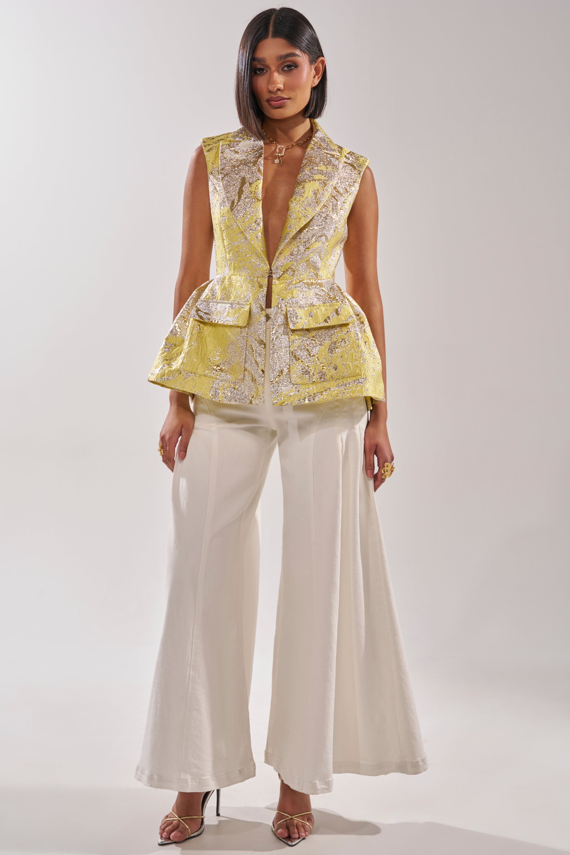 A woman faces forward in the NAOMI BLAZER VEST IN BROCADE, a yellow metallic sleeveless blazer with large front pockets, styled with wide-leg white pants, straight dark hair, gold jewelry, and open-toed heels against a plain background. 4