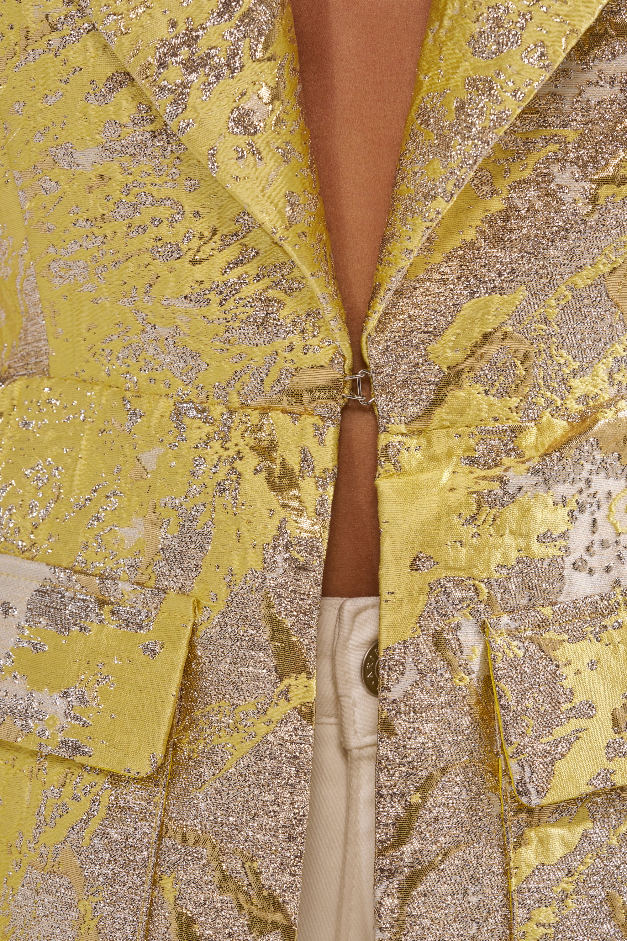 Close-up of the NAOMI BLAZER VEST IN BROCADE, a sleeveless yellow metallic brocade piece fastened with a hook and worn open at the chest, revealing beige or cream pants underneath. 5