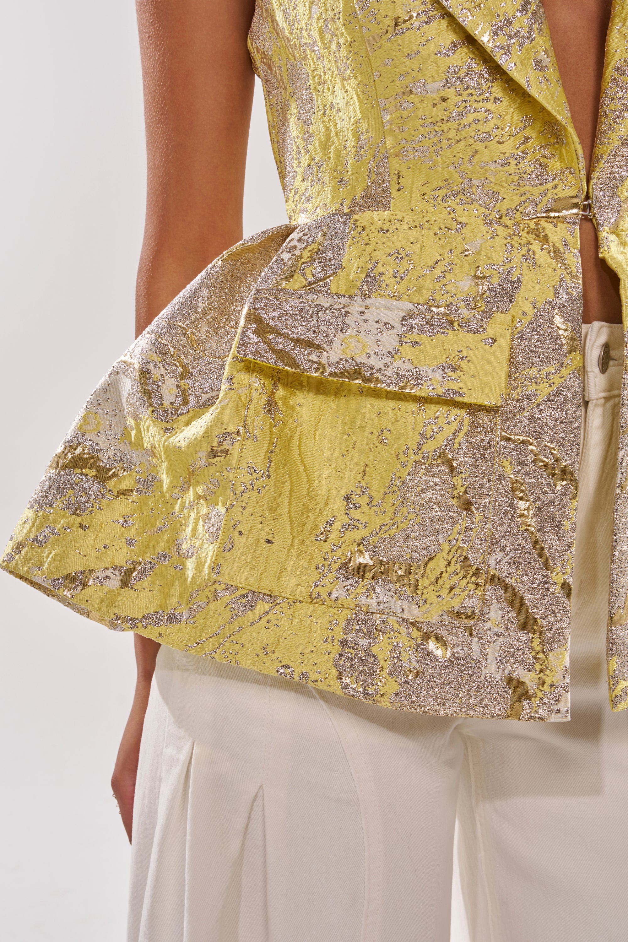 A close-up of a person in white pants and the NAOMI BLAZER VEST IN BROCADE—yellow metallic, sleeveless, with flared sides, large front pockets, and a partially open front that highlights its bold texture and modern silhouette. 6