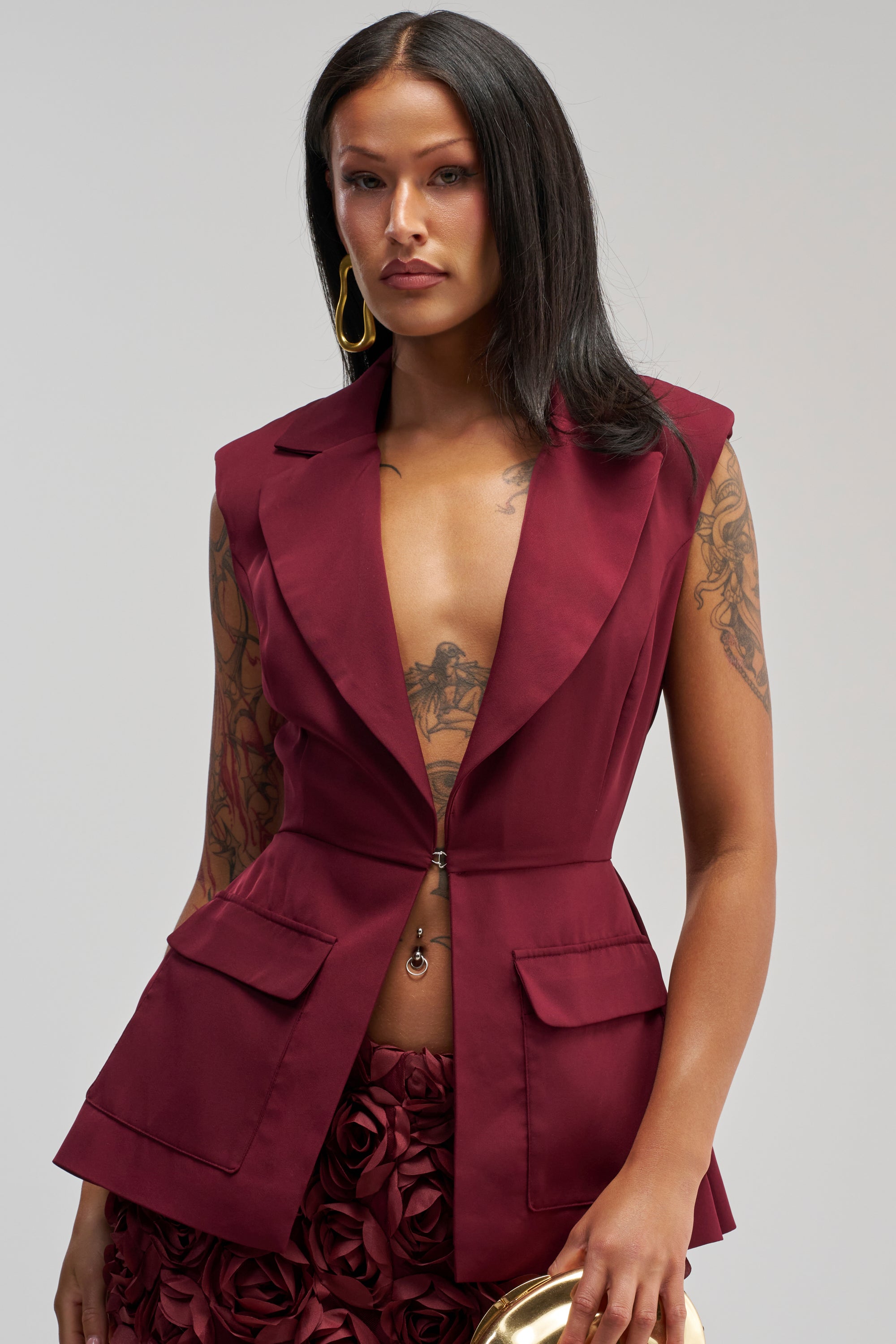 A woman with long dark hair showcases a sharp officewear ensemble, wearing the NAOMI BLAZER VEST IN BURGUNDY— a sleeveless blazer with front pockets and a gold clasp—paired with a textured skirt. Her tattoos are visible on her arms and chest as she holds a gold clutch.