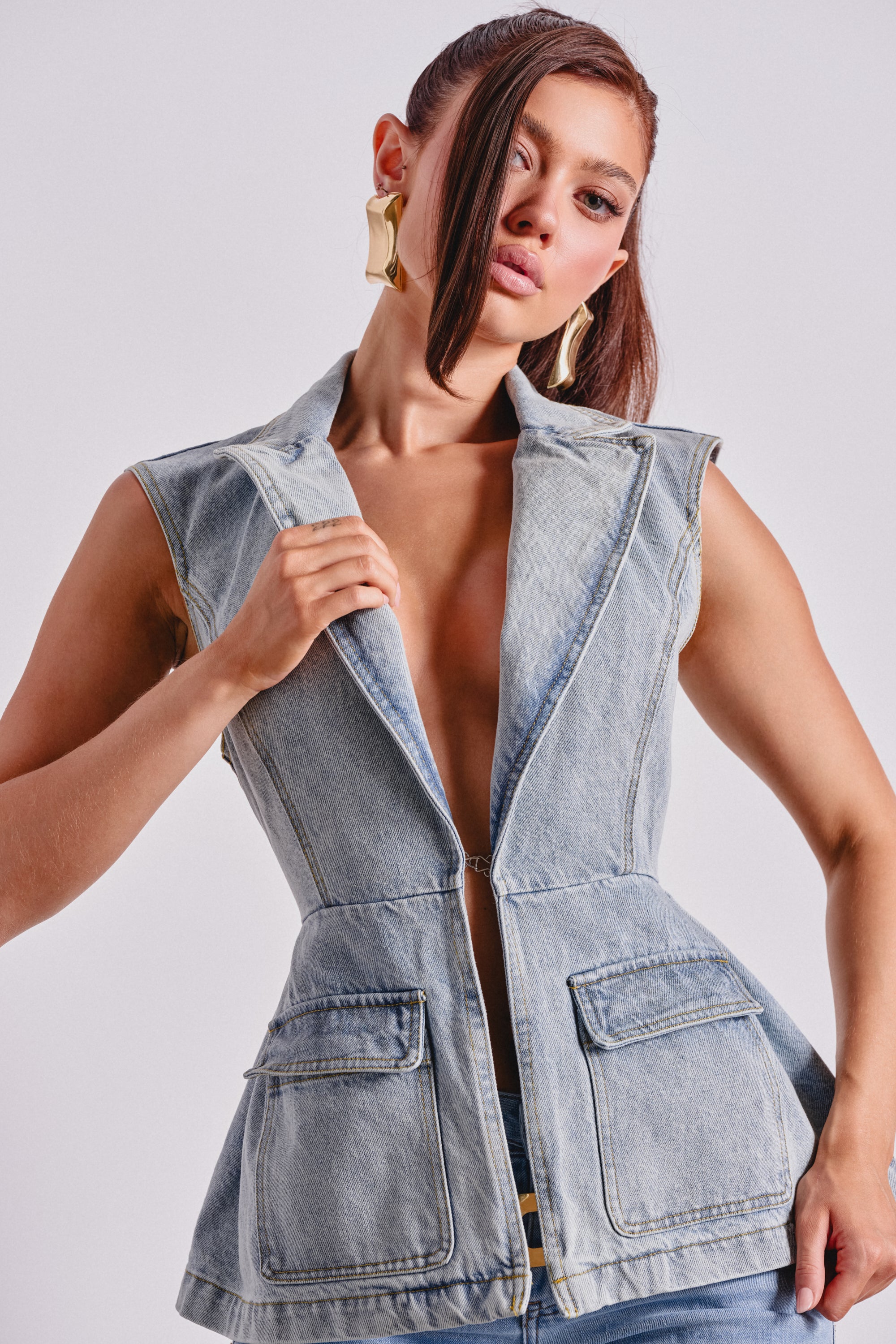 A woman poses confidently against a plain background wearing the SCOUT DENIM BLAZER VEST featuring large front pockets and gold earrings. Her hair is styled with a strand falling over one eye. 0