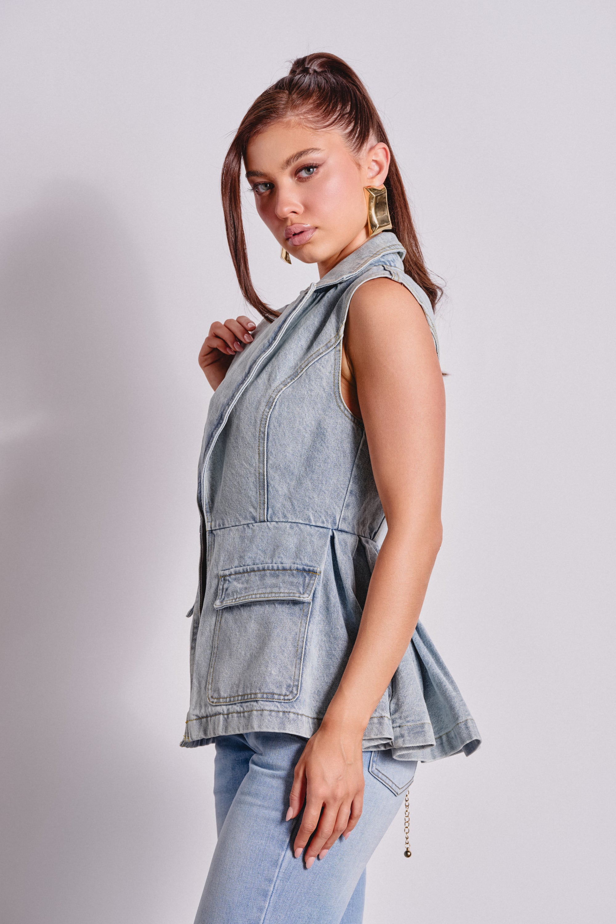 A woman with long brown hair in a high ponytail poses sideways, wearing the SCOUT DENIM BLAZER VEST and matching jeans, accessorized with gold hoop earrings, against a plain light background. 3