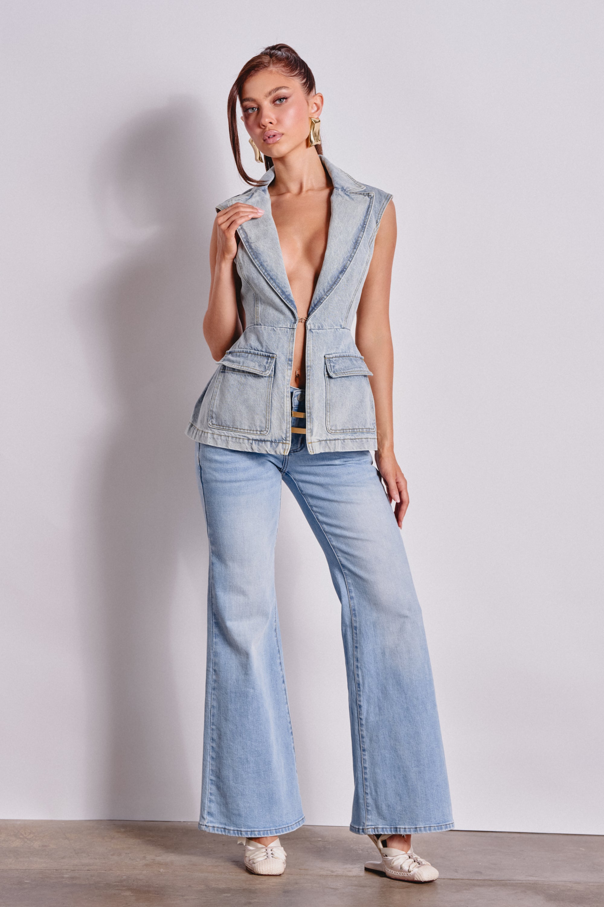 A woman poses against a plain background wearing the SCOUT DENIM BLAZER VEST, styled sleeveless with a deep V-neck, paired with wide-leg light wash denim jeans, white shoes, and statement earrings. One hand rests on her collar. 6