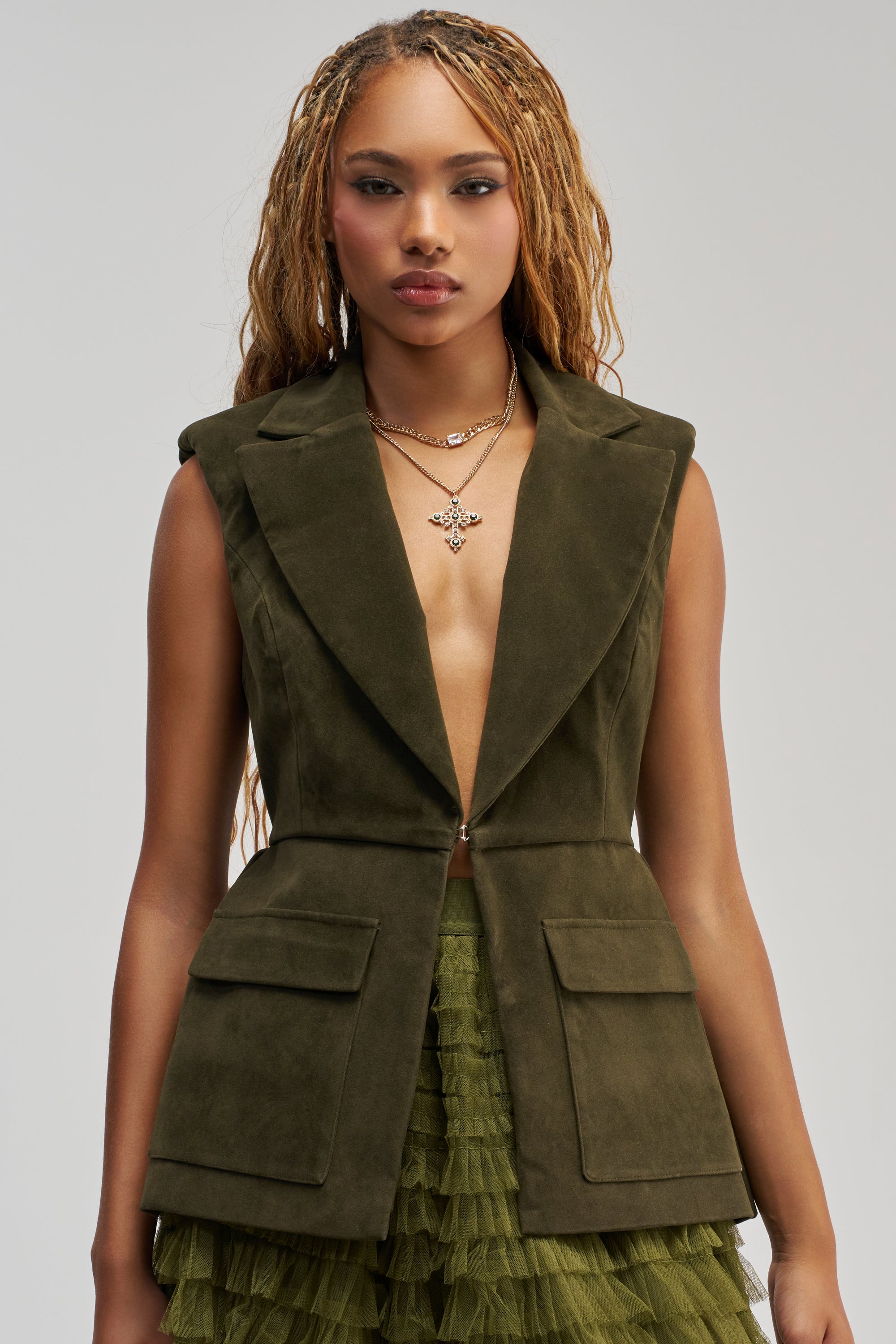 Wearing the NAOMI BLAZER VEST IN SUEDE—a sleeveless olive green blazer with large pockets and a chic peplum hem—a woman with long, braided hair pairs it with a layered tulle skirt. She accessorizes with layered necklaces, one featuring a cross pendant, beautifully capturing sophisticated business casual style as she stands against a neutral background.