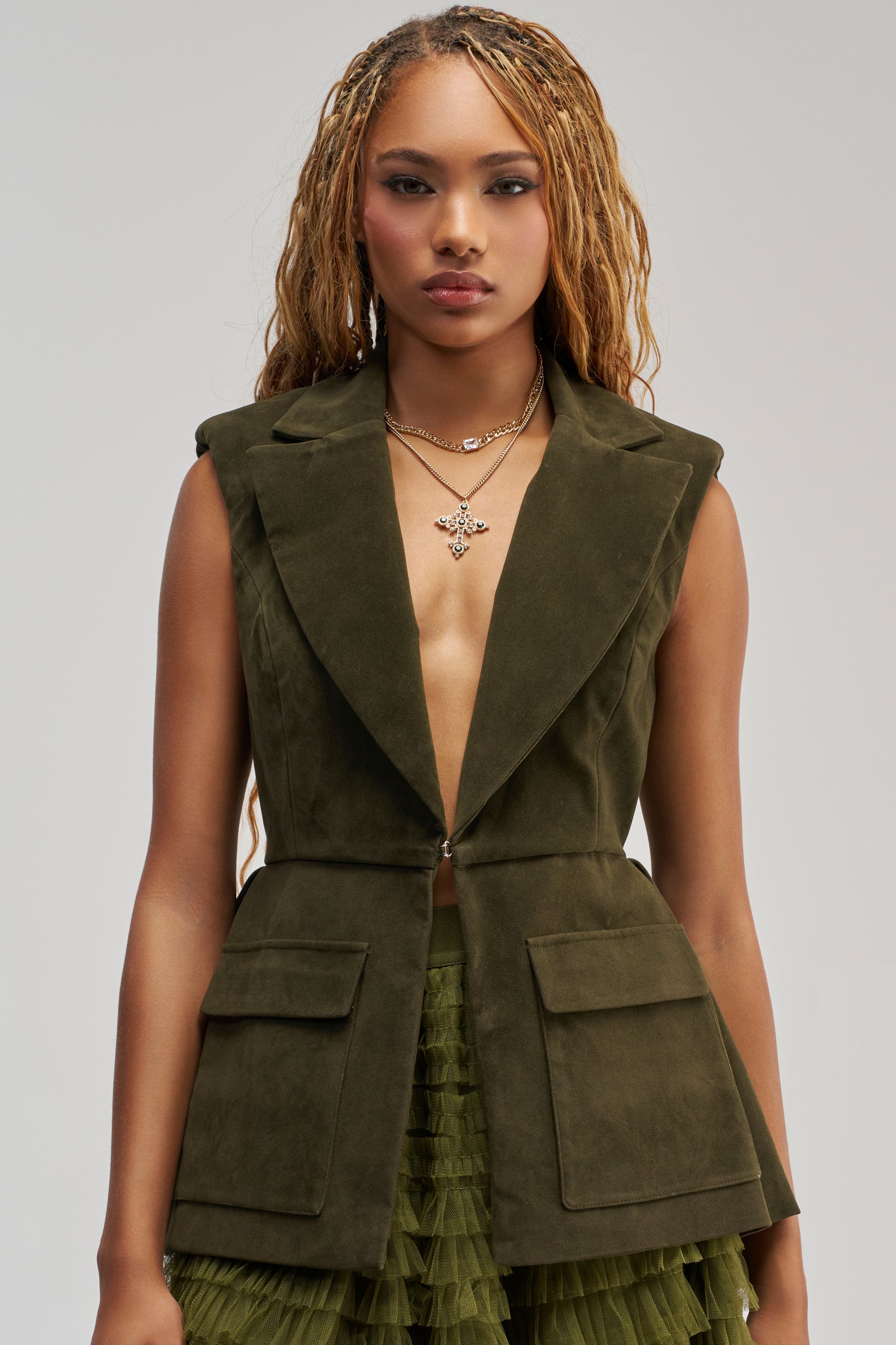 A woman with long braids wears the NAOMI BLAZER VEST IN SUEDE, an olive green sleeveless blazer vest with large lapels and flap pockets. She pairs it with a layered green skirt that features a subtle peplum hemline. With a neutral expression and layered necklaces, she creates a polished business casual look.