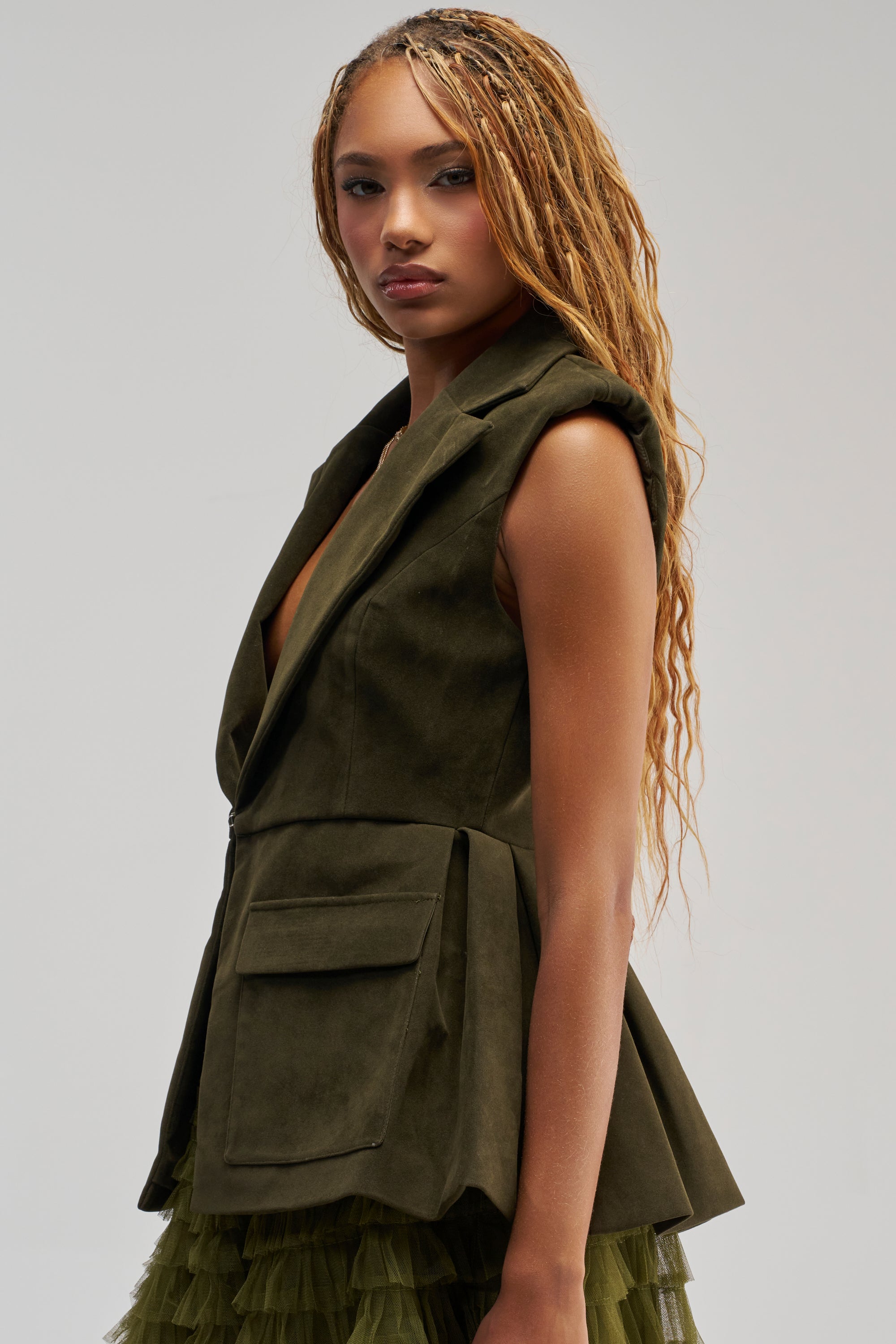 A woman with long, braided hair wears the NAOMI BLAZER VEST IN SUEDE, a green suede vest featuring large front pockets, paired with a layered tulle skirt with a subtle peplum hemline, as she poses against a plain, light background.