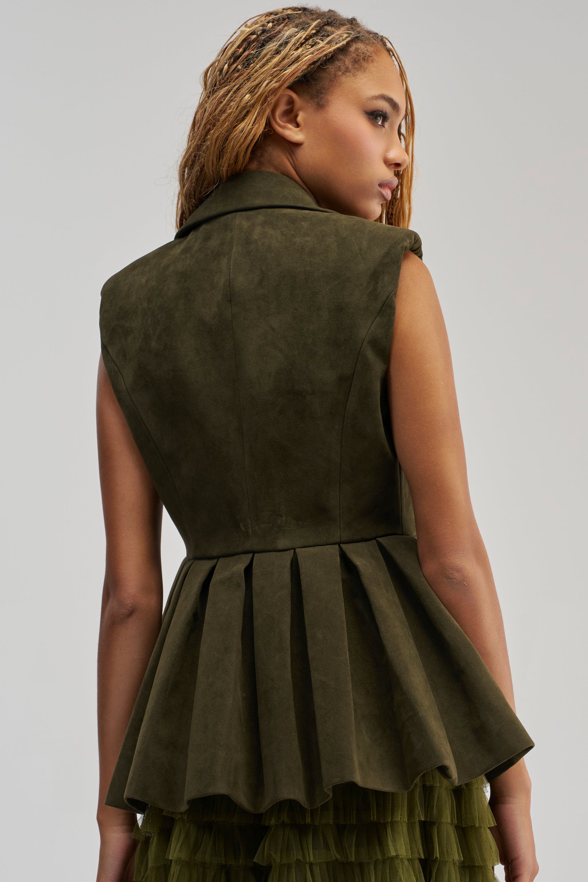 Wearing the NAOMI BLAZER VEST IN SUEDE, a person with braided hair is seen from the back against a plain light background. The sleeveless, dark green blazer vest features a pleated peplum hemline and is paired with a green ruffled skirt, offering an elegant twist on business casual attire.