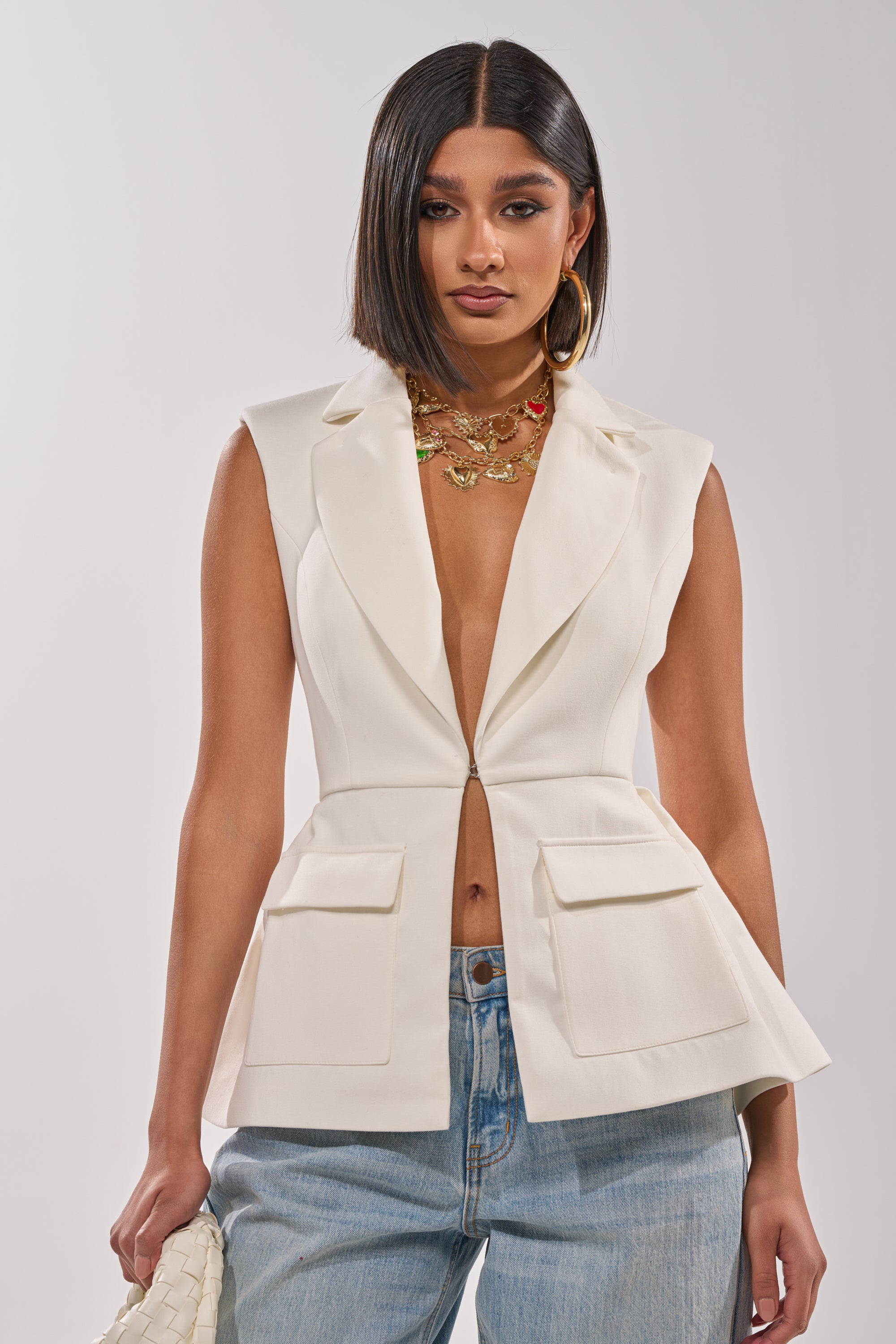 A woman with straight, shoulder-length dark hair wears the NAOMI BLAZER VEST IN WHITE with large pockets and light blue jeans. She accessorizes with layered gold necklaces, hoop earrings, and a white bag. 0
