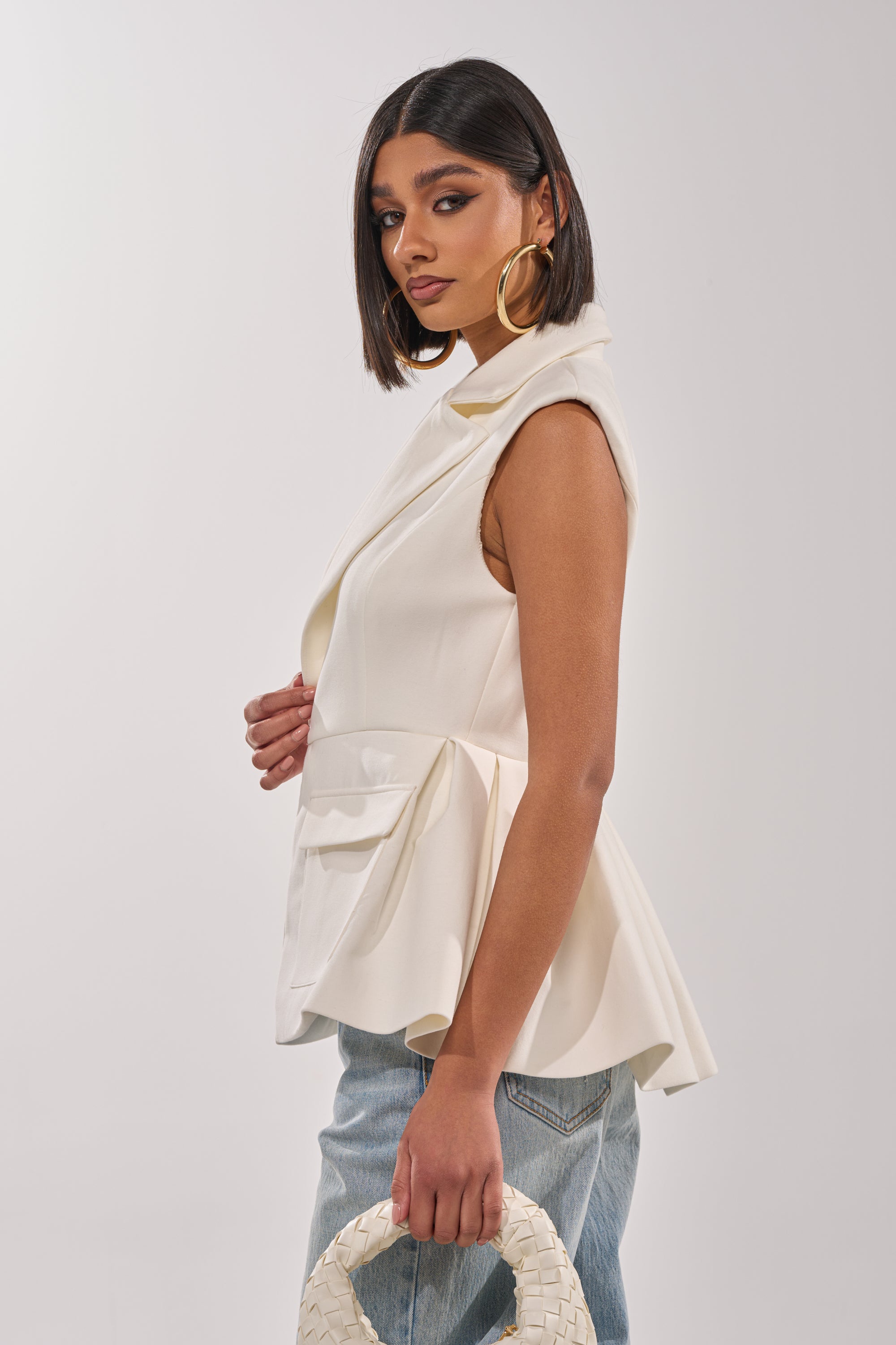 A woman with straight dark hair and gold hoop earrings wears the NAOMI BLAZER VEST IN WHITE with light blue jeans, holding a white woven handbag, posing against a plain light background. 2