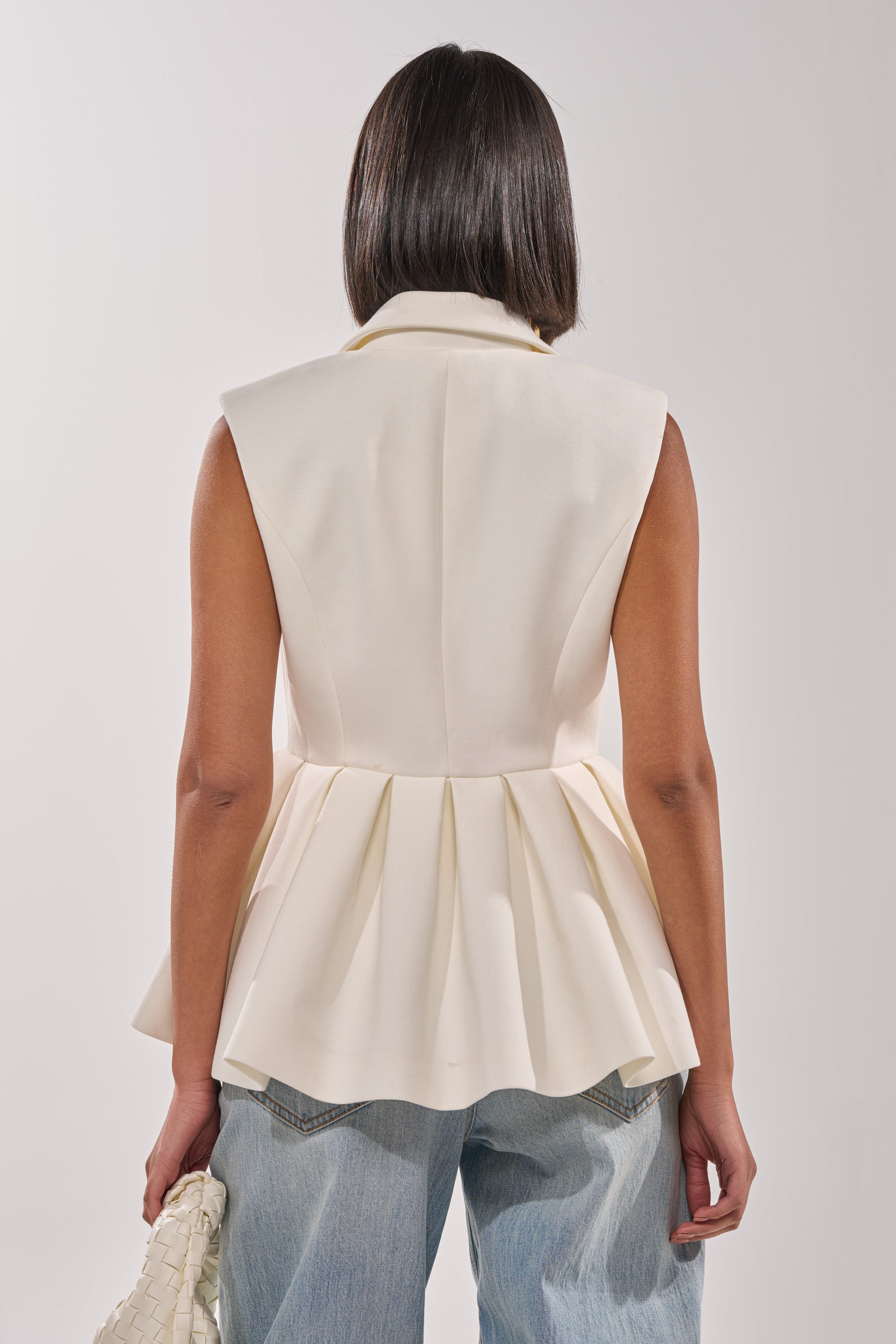 A woman, seen from behind, wears the NAOMI BLAZER VEST IN WHITE with blue jeans and holds a white woven handbag in her left hand against a plain light background for a chic business-casual look. 3