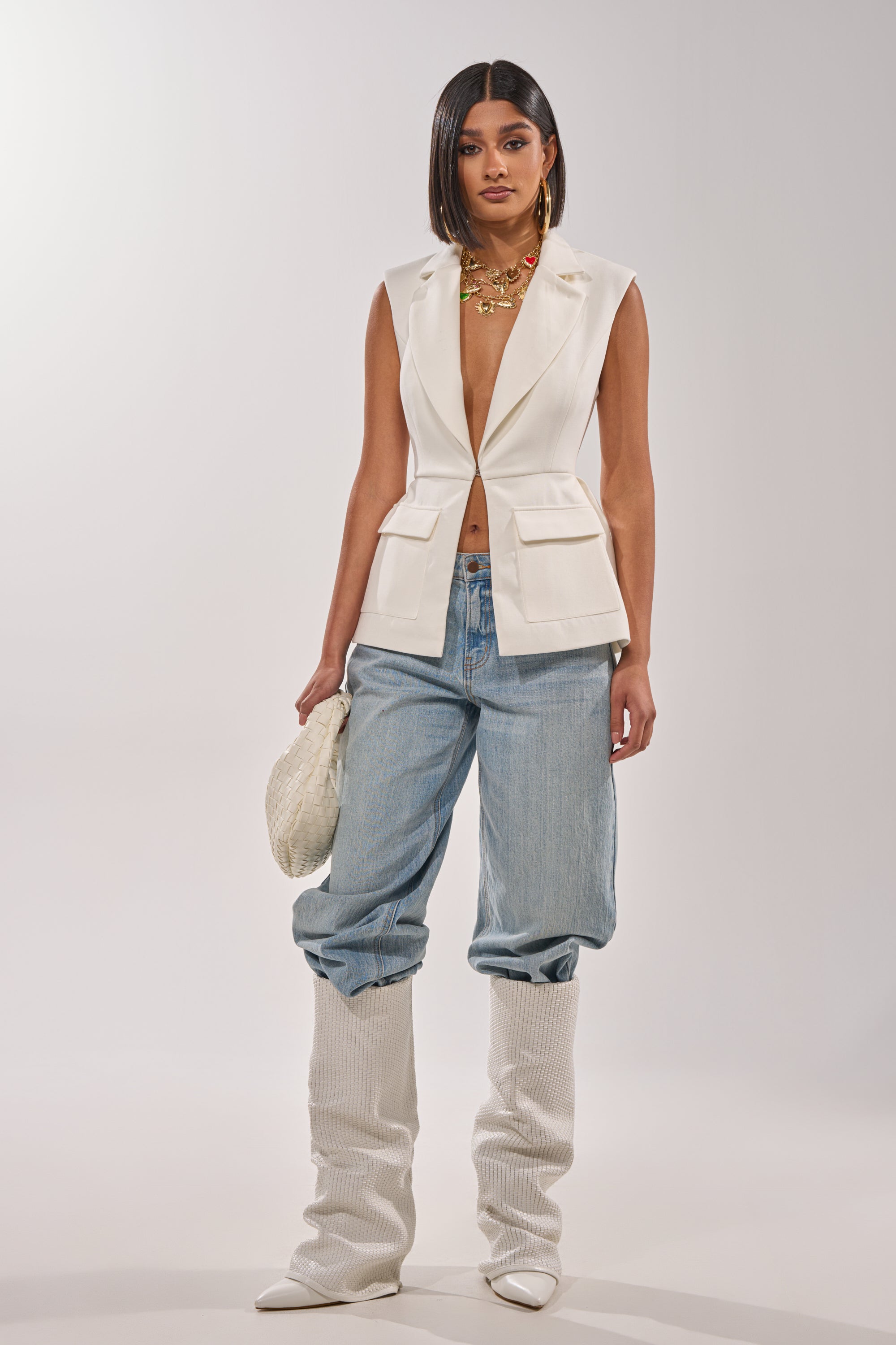 A woman wears the NAOMI BLAZER VEST IN WHITE styled with light blue baggy jeans, tall white textured boots, a white woven clutch, and layered gold necklaces, creating a chic business-casual look against a plain light background. 4