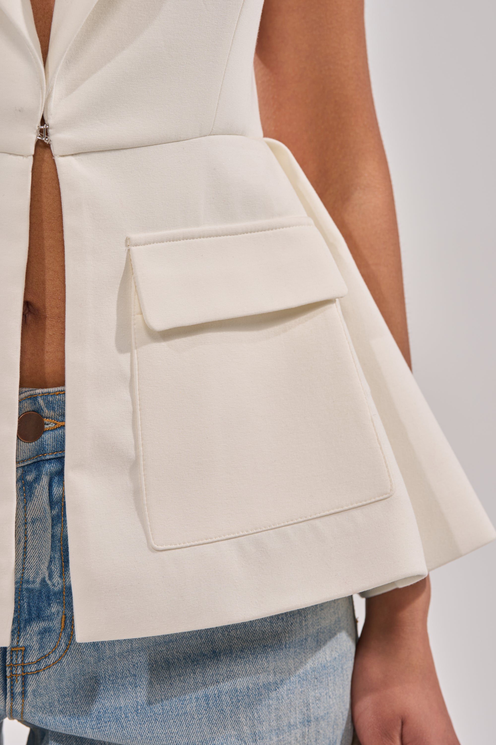 A close-up of a person wearing the NAOMI BLAZER VEST IN WHITE with a large front pocket, styled open at the midriff and paired with light blue denim jeans for an effortless business-casual look. 7