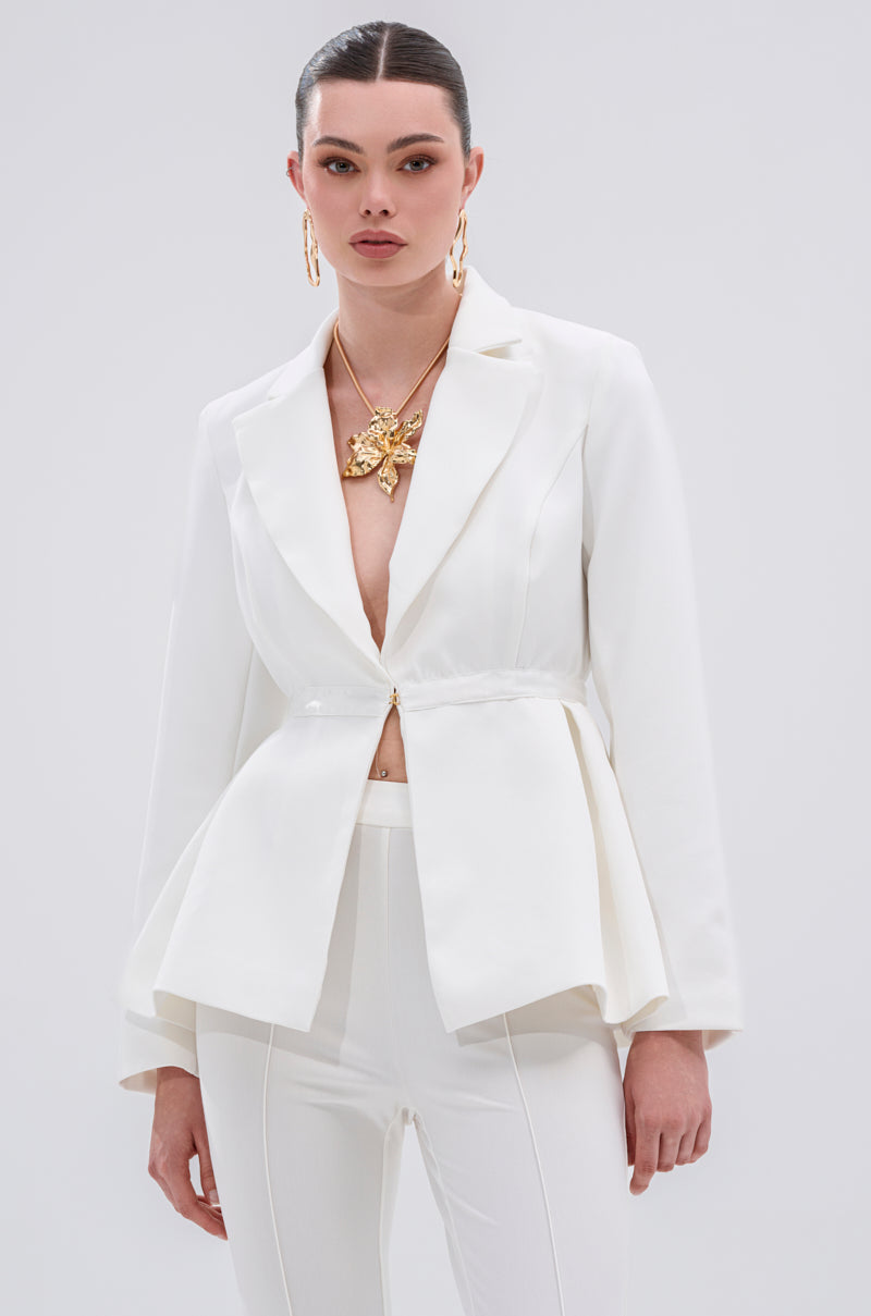 A woman models the CANARY PEPLUM BLAZER IN WHITE—featuring a deep V-neck and gold button details—against a light background, styled with bold gold jewelry, slicked-back hair, and a neutral expression.
