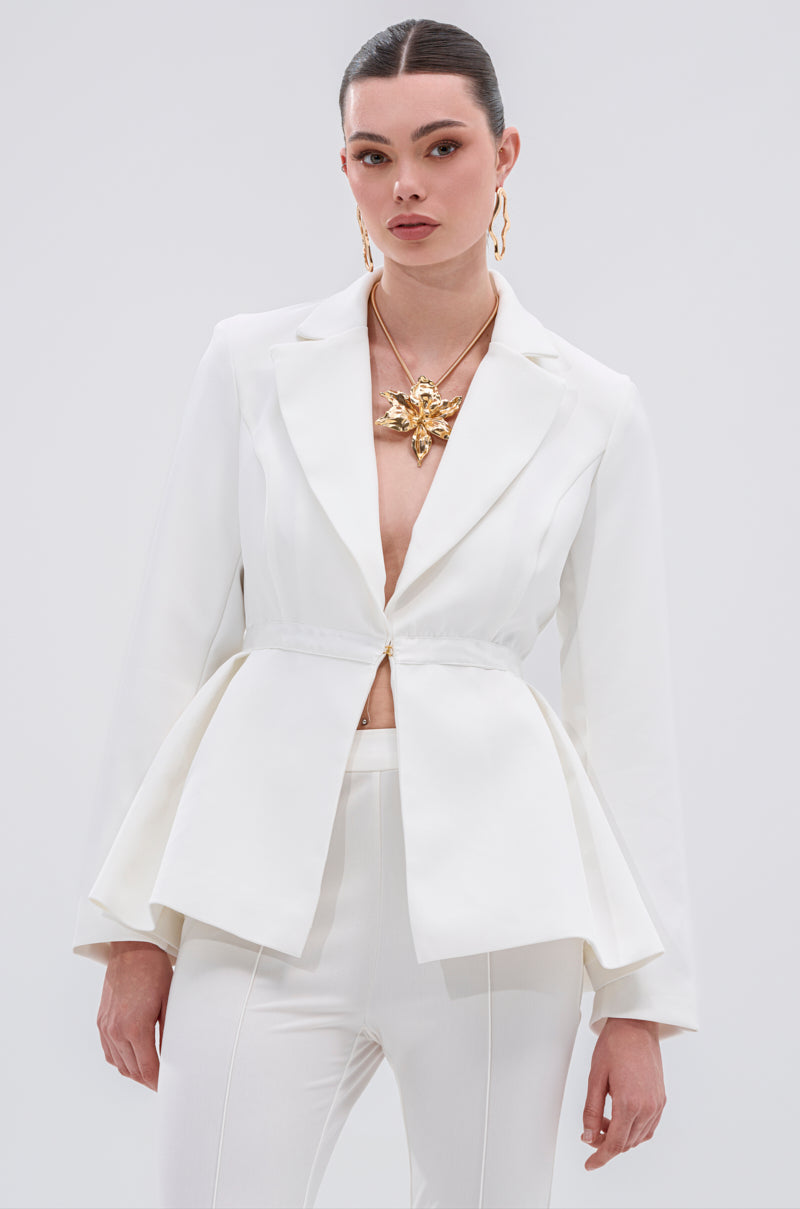 A woman stands against a plain background wearing the CANARY PEPLUM BLAZER IN WHITE, featuring gold metallic buttons and matching pants. Her look is completed with gold earrings, a statement necklace, and slicked-back dark hair.