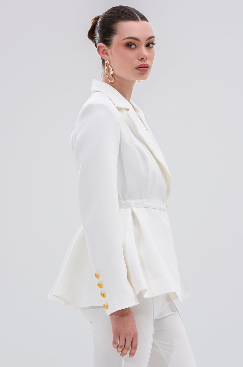 A woman with dark hair in a sleek bun models the CANARY PEPLUM BLAZER IN WHITE with gold button detailing, paired with matching white pants and statement earrings, set against a plain light background.