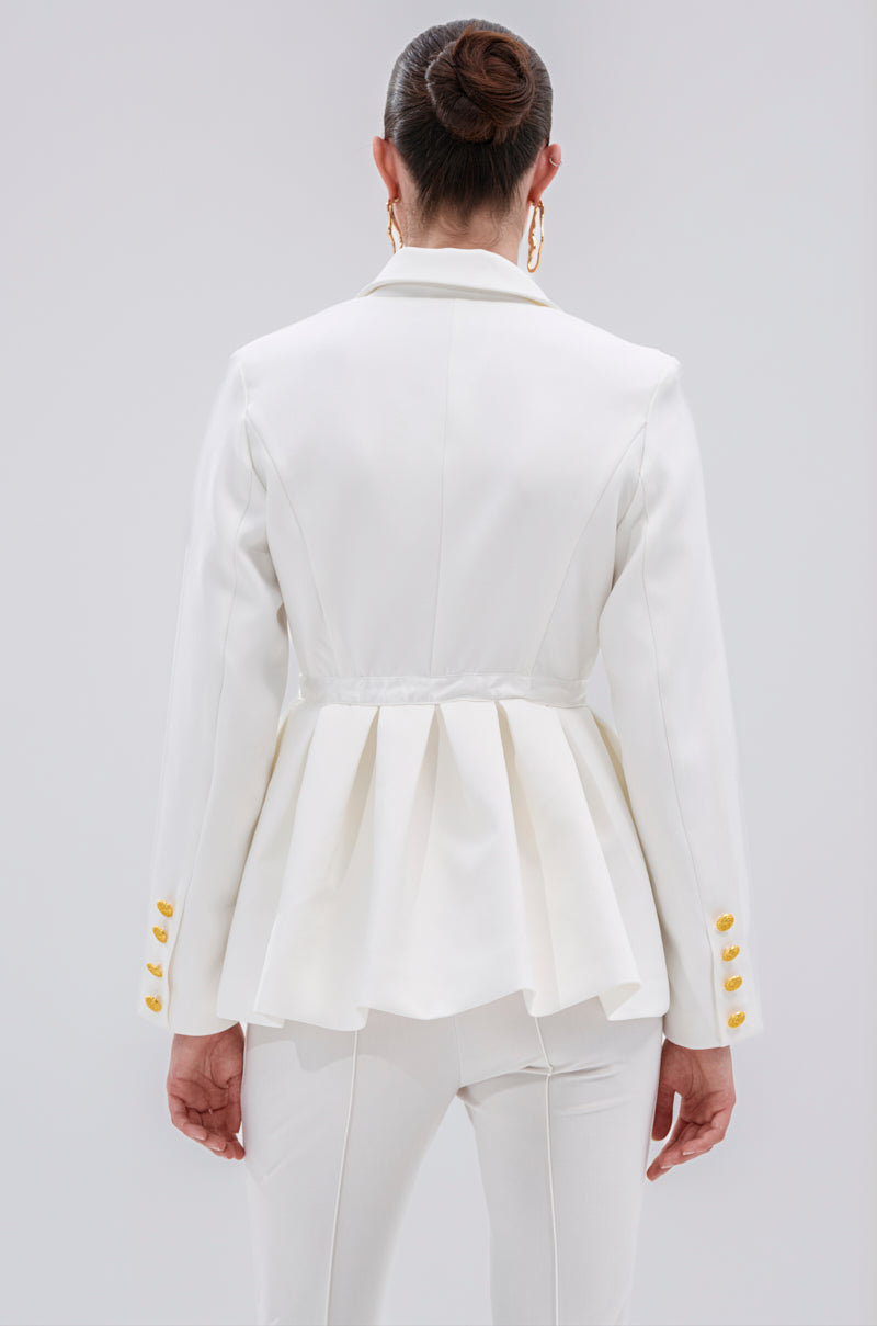 A woman with dark hair in a bun and gold hoop earrings stands facing away, wearing the CANARY PEPLUM BLAZER IN WHITE—featuring gold metallic buttons and a pleated waist—paired with matching white pants.