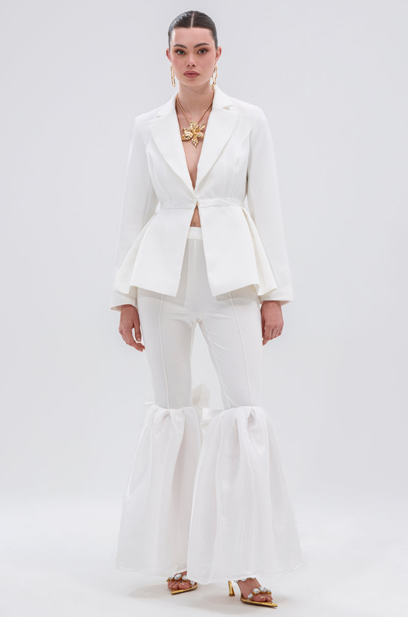 A woman poses against a plain backdrop in the CANARY PEPLUM BLAZER IN WHITE, featuring gold metallic buttons. She pairs it with matching flared pants, accessorized with gold jewelry, slicked-back hair, and strappy heels.