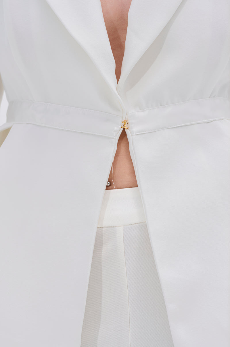 Close-up of a person wearing the CANARY PEPLUM BLAZER IN WHITE, fastened by a single gold metallic button and paired with high-waisted white pants, revealing a hint of bare skin underneath.