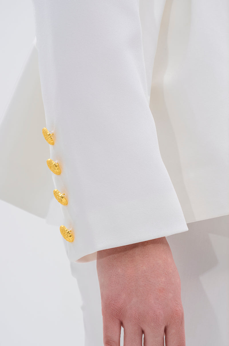 A close-up of an arm wearing the CANARY PEPLUM BLAZER IN WHITE, showcasing four gold buttons on the sleeve. The hand rests relaxed by their side, highlighting this elegant tailored statement piece.