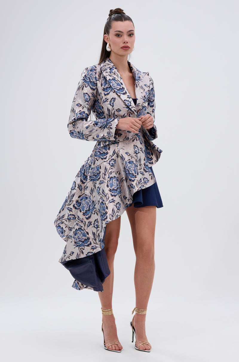 Woman wearing the CHARLOTTE ASYMMETRICAL BLAZER in blue and beige floral jacquard with a ruffled hem, paired with a navy underskirt, hoop earrings, and gold high-heeled sandals against a plain background.