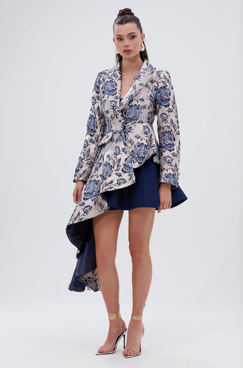 A woman wears the CHARLOTTE ASYMMETRICAL BLAZER in floral metallic jacquard with a long draped side, paired with a short navy skirt, gold sandals, and her dark hair styled in a sleek high ponytail.