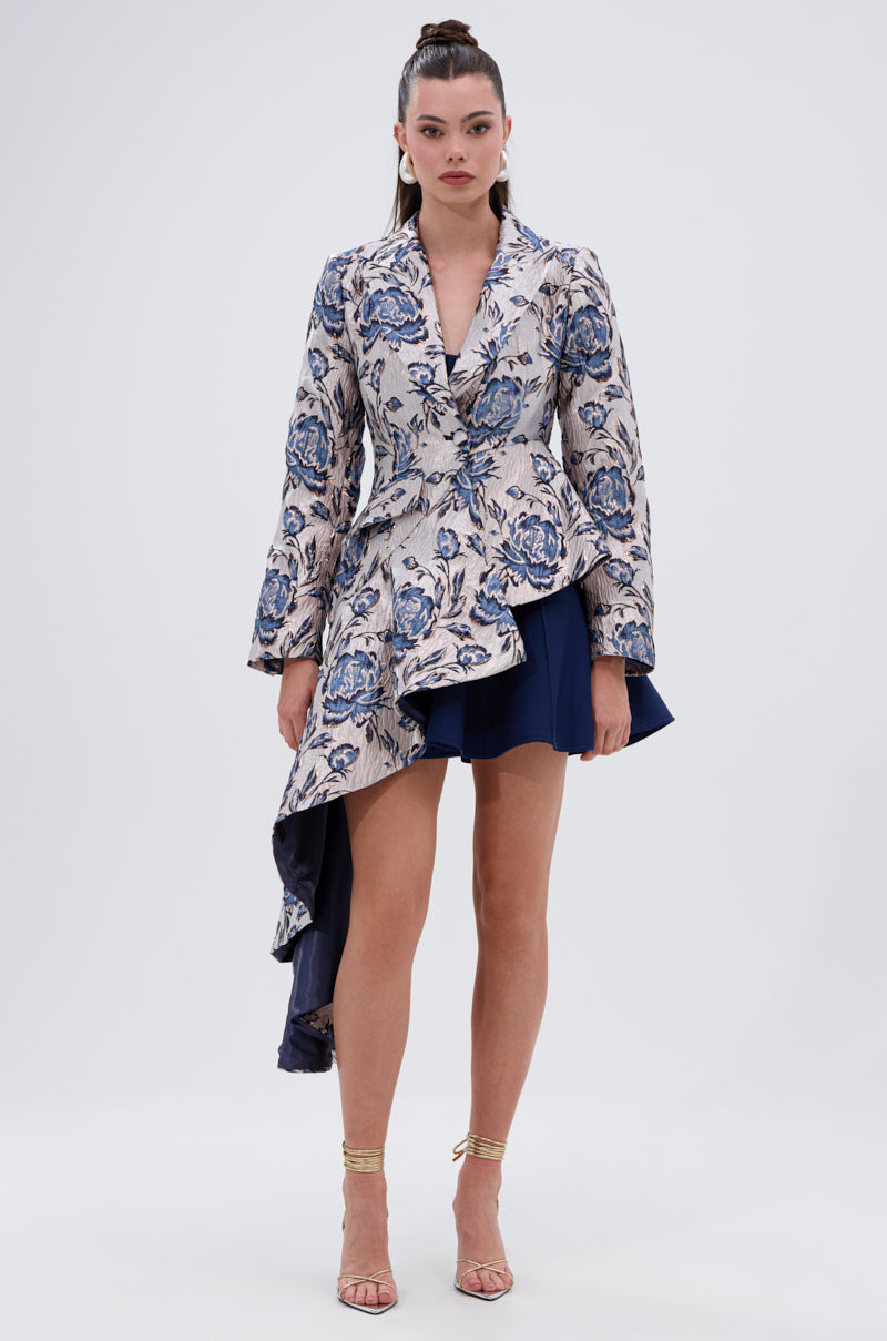 A woman wears the CHARLOTTE ASYMMETRICAL BLAZER with a floral pattern and dramatic hem, styled with a navy skirt and gold heels. Her hair is in a high ponytail.