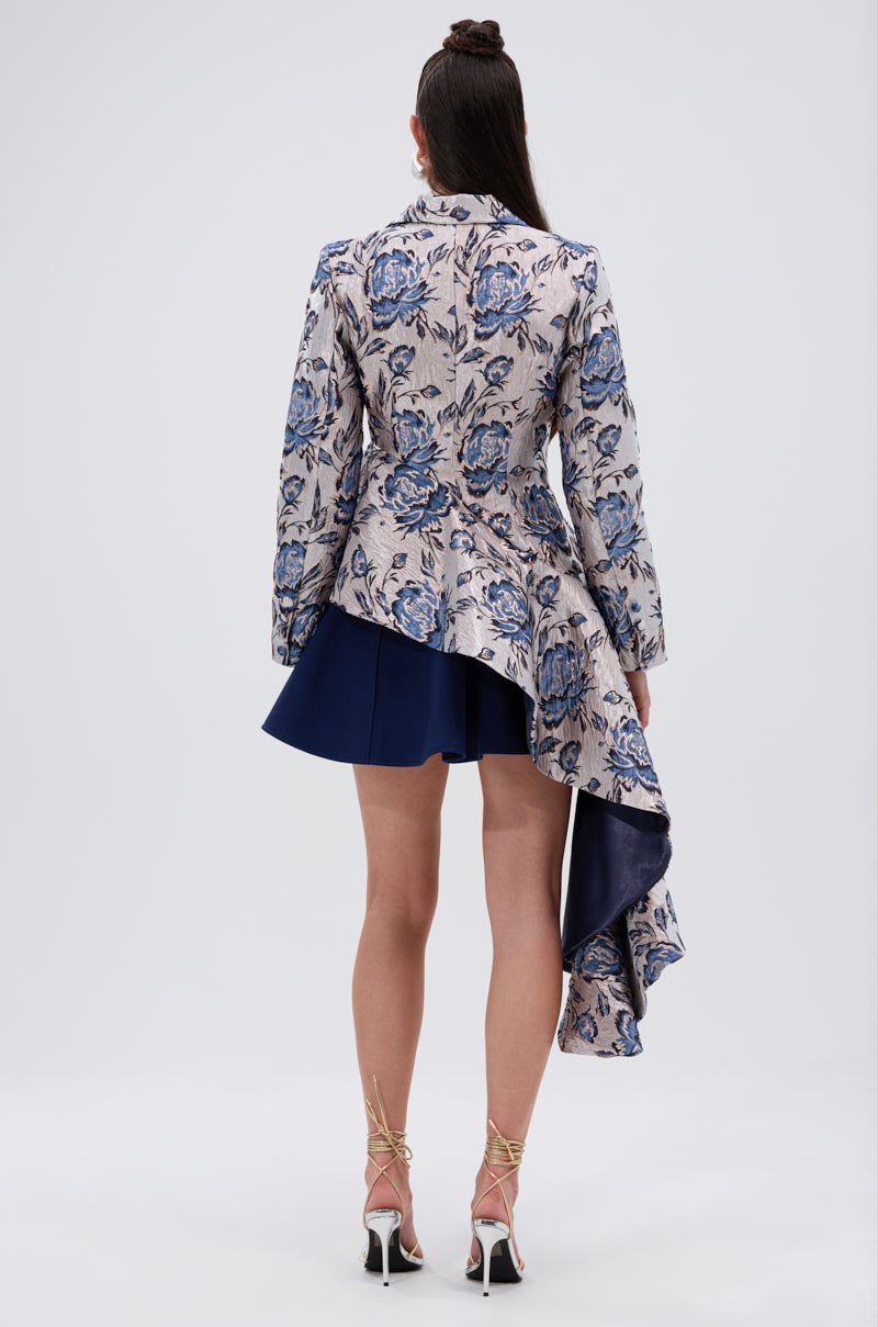 A woman with long, half-up brown hair wears the CHARLOTTE ASYMMETRICAL BLAZER featuring a floral peplum silhouette over a short navy skirt, paired with high-heeled sandals.