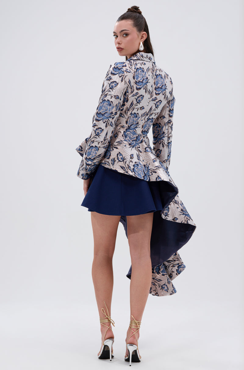 A woman models the CHARLOTTE ASYMMETRICAL BLAZER, featuring a floral pattern with blue accents, styled with a short navy skirt and strappy high heels against a plain light background.