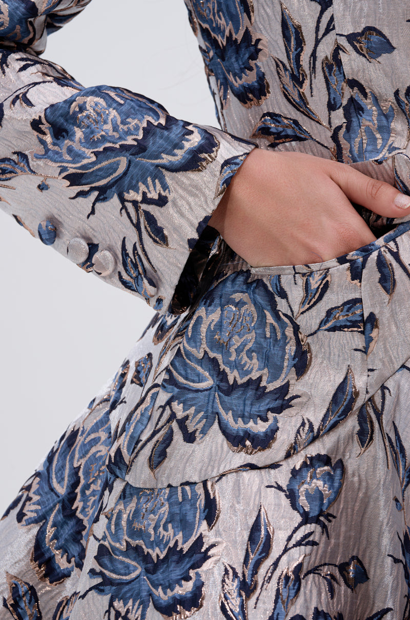 A close-up of a person wearing the CHARLOTTE ASYMMETRICAL BLAZER in silver and blue metallic jacquard, featuring bold blue florals, an asymmetrical cut, and one hand tucked into the pocket.