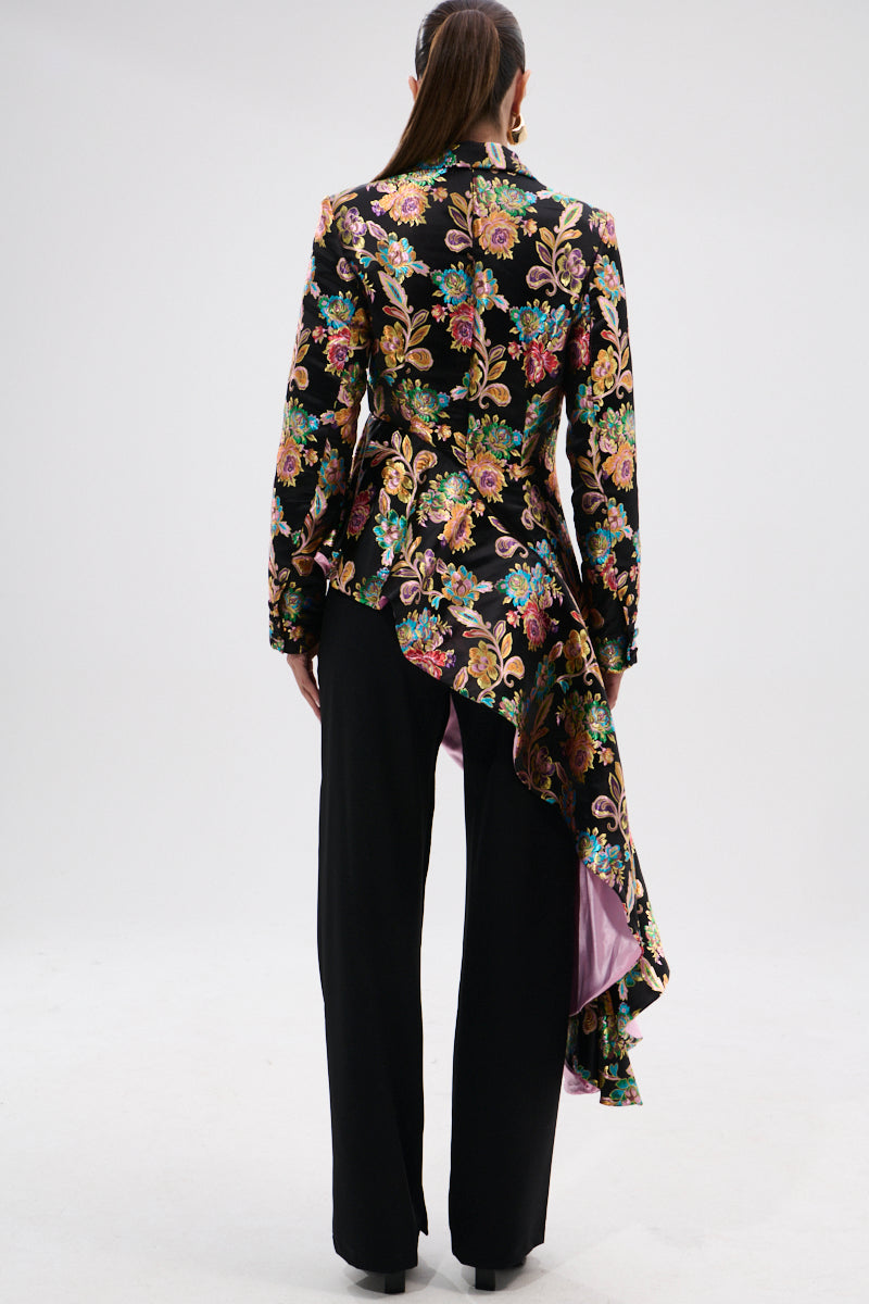 A person stands facing away, wearing black pants and the STEPPING OUT ASYMMETRICAL BLAZER in floral brocade with a purple-lined cascading hem. Their hair is styled in a sleek ponytail, set against a plain white background.