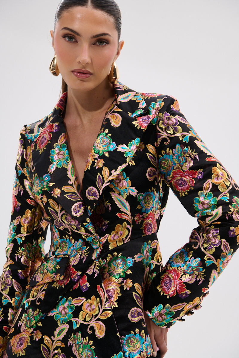A woman with slicked-back hair and gold hoop earrings poses confidently, hand on hip, in the STEPPING OUT ASYMMETRICAL BLAZER—black with vibrant colorful floral embroidery—against a plain background.