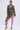A woman stands confidently in the STEPPING OUT ASYMMETRICAL BLAZER, featuring a floral brocade design, over a pink lace jumpsuit and matching heels—an outfit blending tradition, vibrant color, modern tailoring, and striking detail.