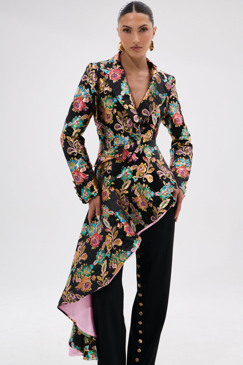 A woman stands confidently in the STEPPING OUT ASYMMETRICAL BLAZER—black floral brocade with a pink-lined peplum hem—paired with black pants featuring gold buttons, set against a plain white background.