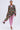A woman models the STEPPING OUT ASYMMETRICAL BLAZER, featuring a black floral brocade design with an asymmetric hem, layered over a pink lace top and matching pants, accessorized with hoop earrings and open-toe heels.