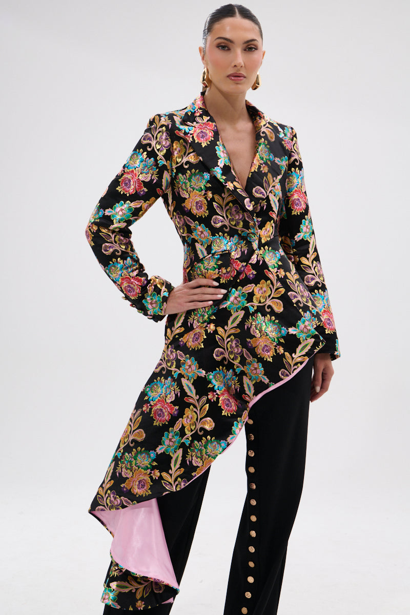 A woman poses in the STEPPING OUT ASYMMETRICAL BLAZER, crafted from vibrant floral brocade with a pink-lined asymmetrical hem, paired with black pants featuring a row of gold buttons down the front.