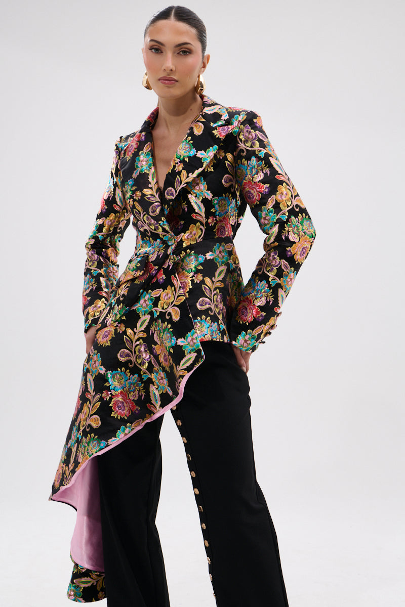 A woman stands confidently in the STEPPING OUT ASYMMETRICAL BLAZER, a black satin jacquard suit with a vibrant floral pattern, pink lining, and matching black pants detailed with gold side buttons. The background is plain white.