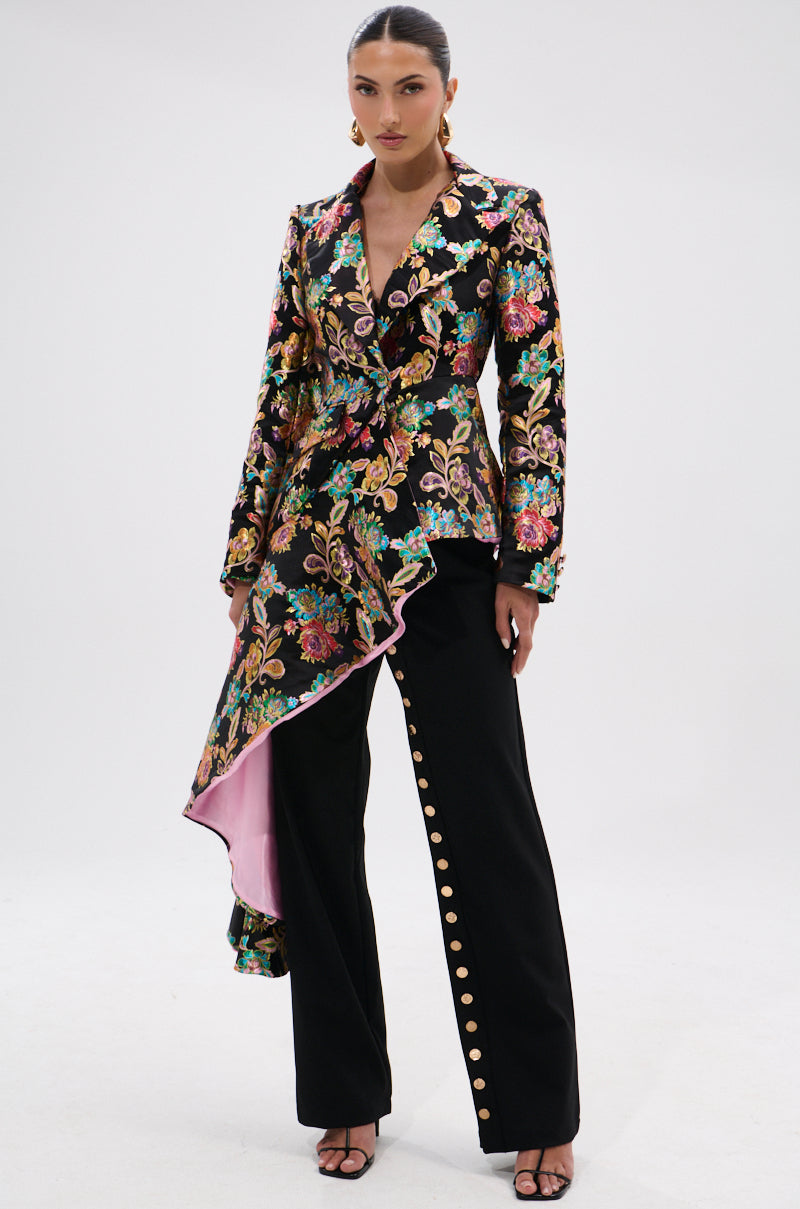 A woman wears the STEPPING OUT ASYMMETRICAL BLAZER, a bold floral brocade piece with a ruffled, pink-lined hem. She pairs it with black pants featuring side gold buttons, open-toe heels, statement earrings, and slicked-back hair.