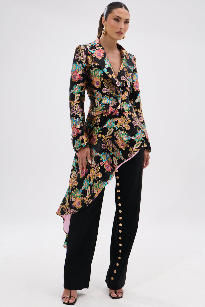 A woman poses in a chic outfit featuring the STEPPING OUT ASYMMETRICAL BLAZER, styled with high-waisted black pants detailed with gold buttons down one leg and black open-toe heels.