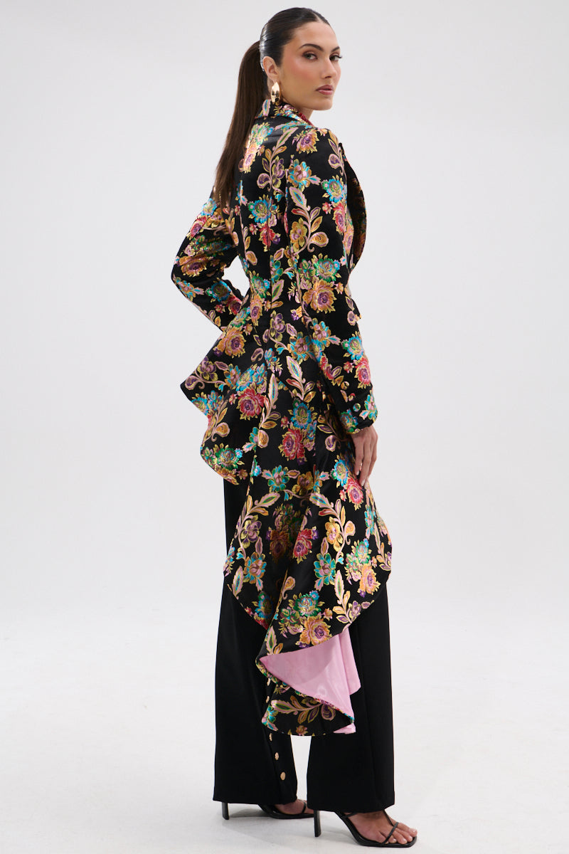 A woman wears the STEPPING OUT ASYMMETRICAL BLAZER with floral brocade and dramatic back ruffle, styled with black wide-leg pants, heeled sandals, bold gold earrings, and a sleek ponytail against a plain background.