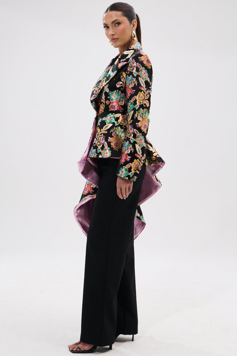A woman stands sideways against a plain white background, wearing the STEPPING OUT ASYMMETRICAL BLAZER with wide-leg black trousers, black heels, her hair pulled back, and large statement earrings.