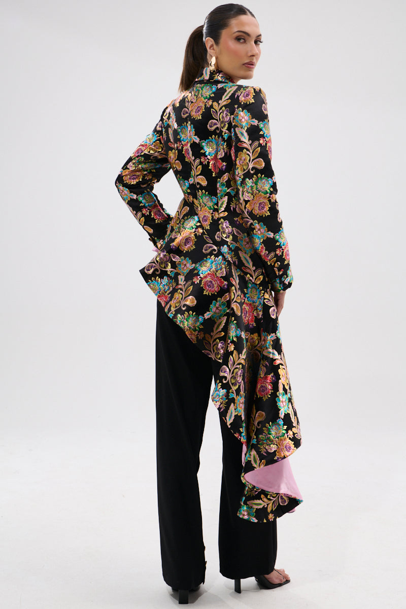 A woman faces away in the STEPPING OUT ASYMMETRICAL BLAZER, a black floral brocade piece with ruffled hem, pink satin jacquard lining showing through. She pairs it with black pants, hoop earrings, heels, and a ponytail.